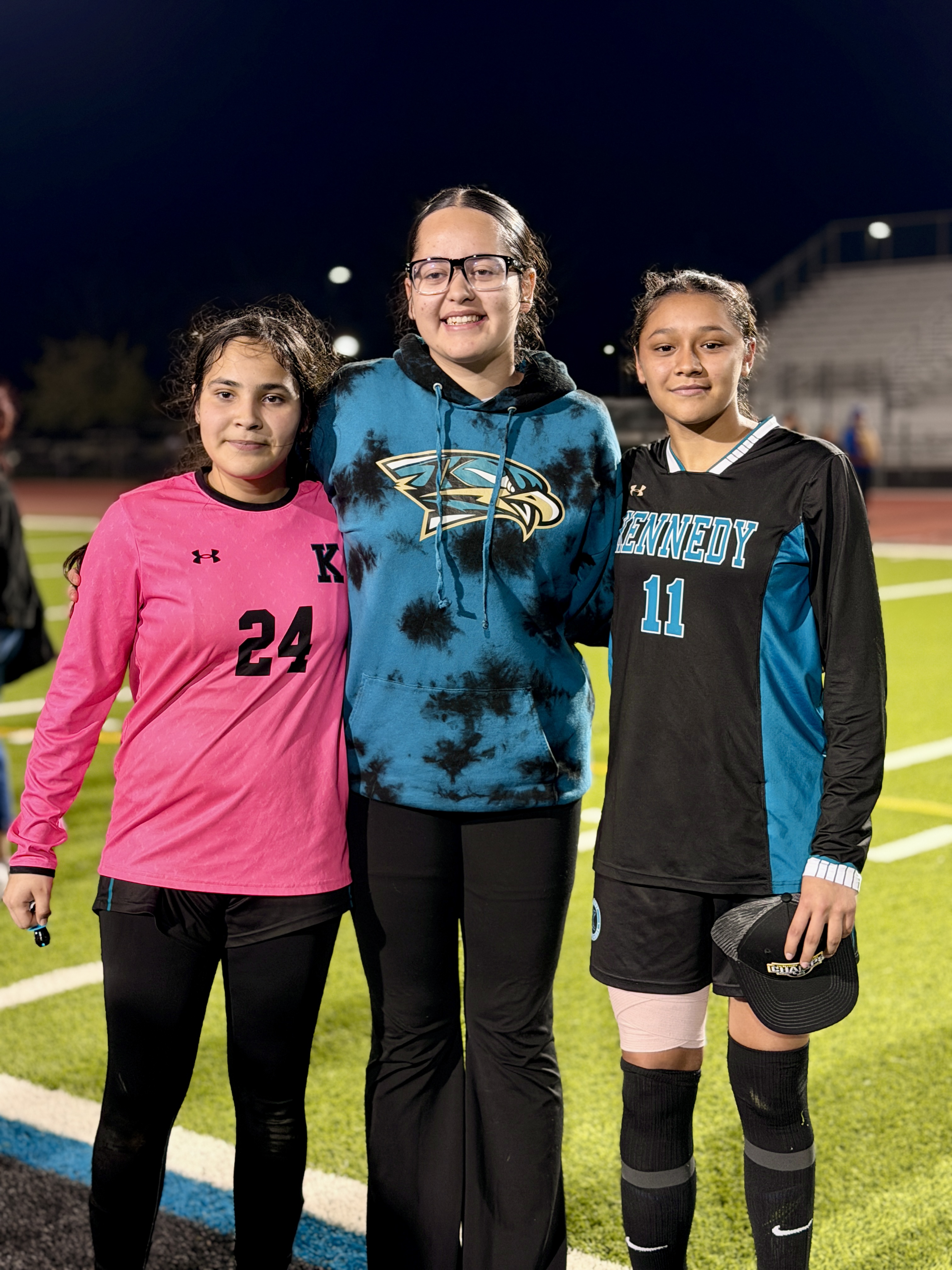 Three young female athletes, wearing sports uniforms, are standing together on a sports field at night.