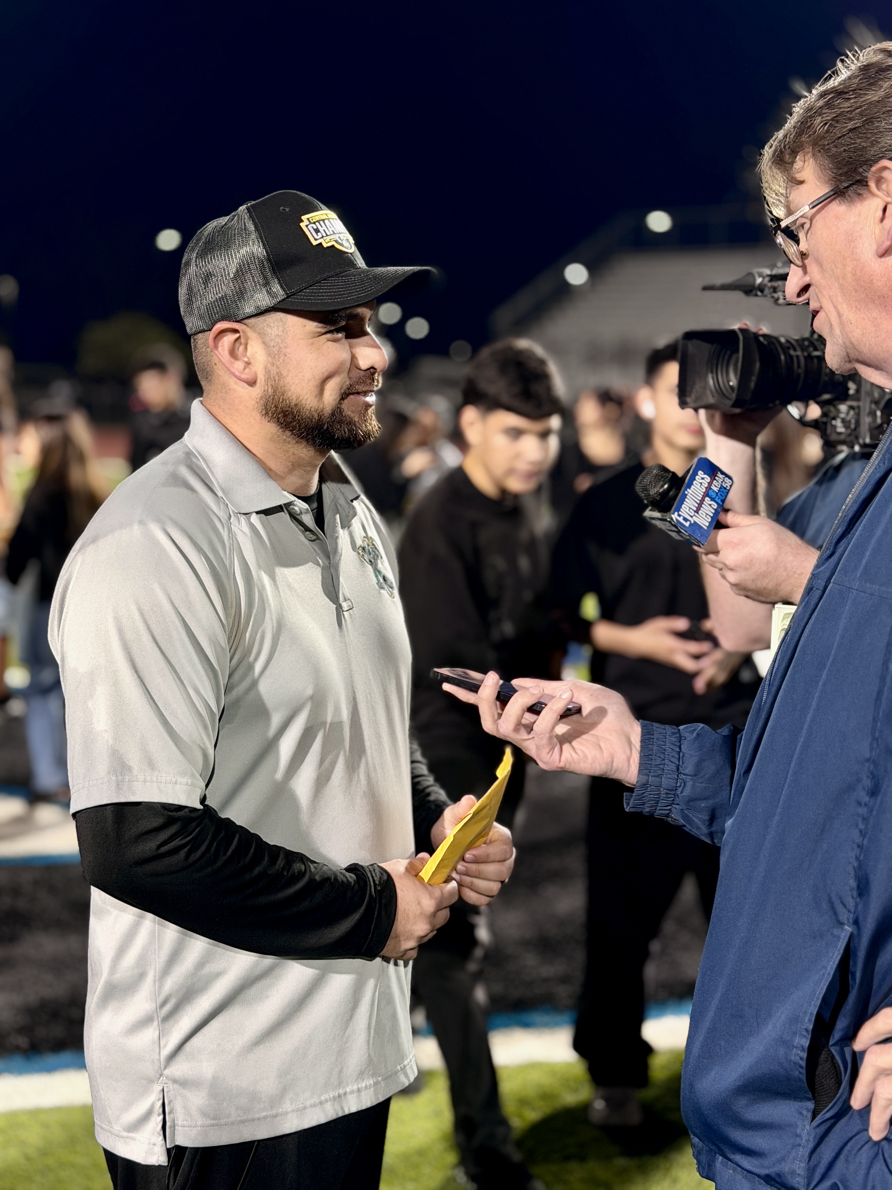 A man in a baseball cap is being interviewed by a reporter holding a microphone in front of a crowd of people at a nighttime event.