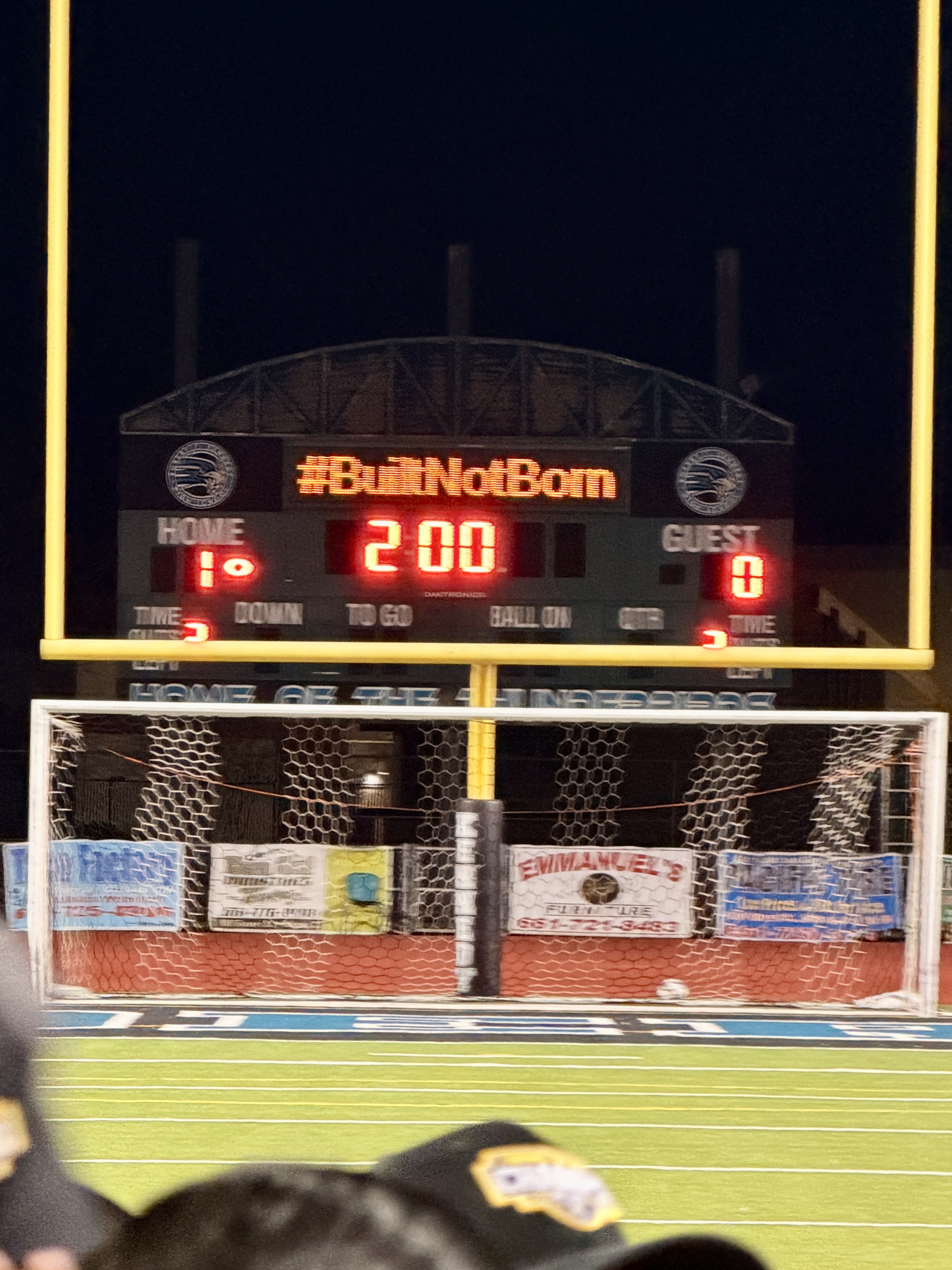 The image shows a scoreboard at a sports stadium, displaying the score of a game with the home team leading 2-00 over the guest team. The scoreboard is surrounded by a metal frame and stands against a dark background, likely the night sky.