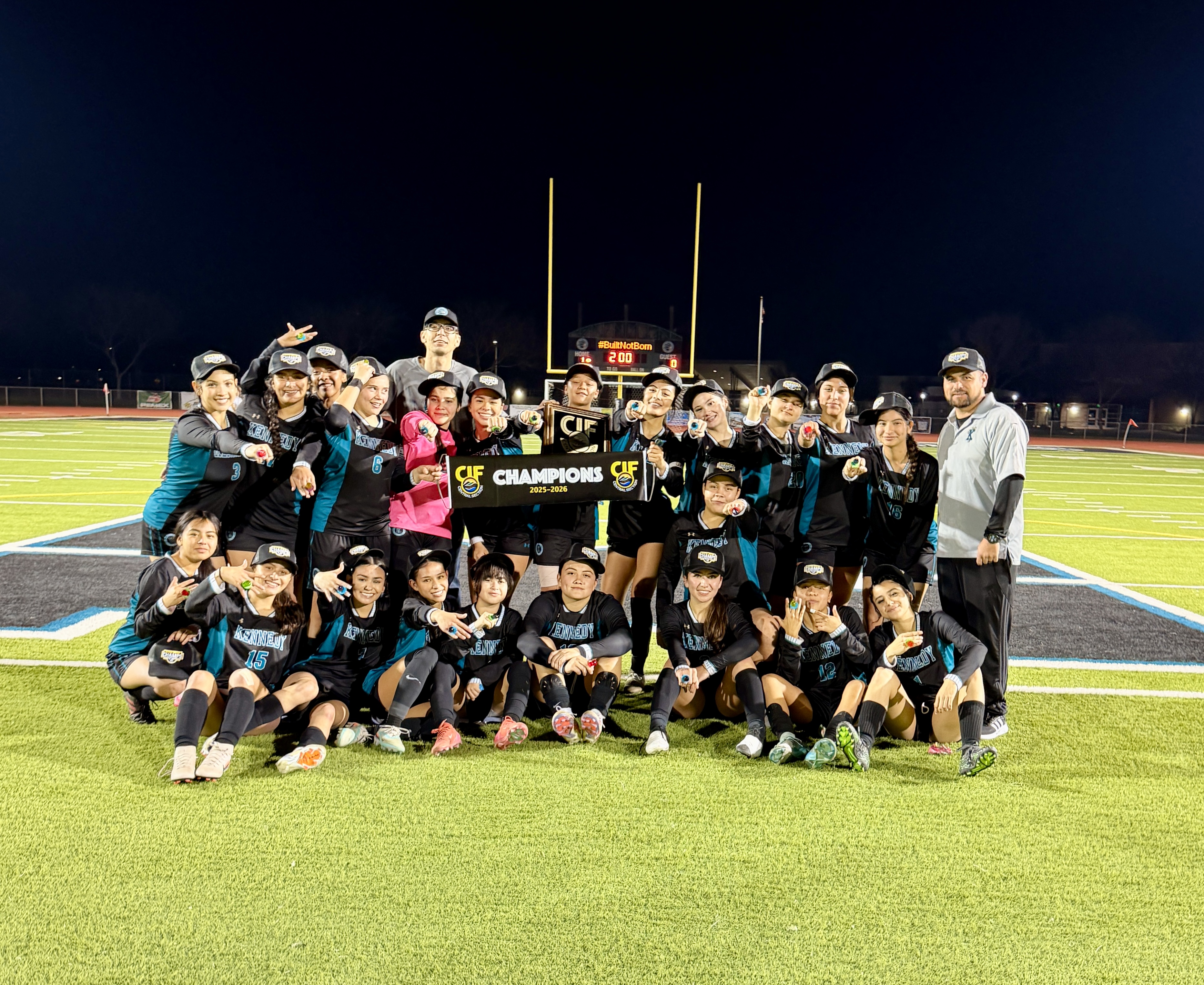 A group of people, likely a sports team, celebrating on a well-lit sports field at night, holding a trophy and wearing matching uniforms.