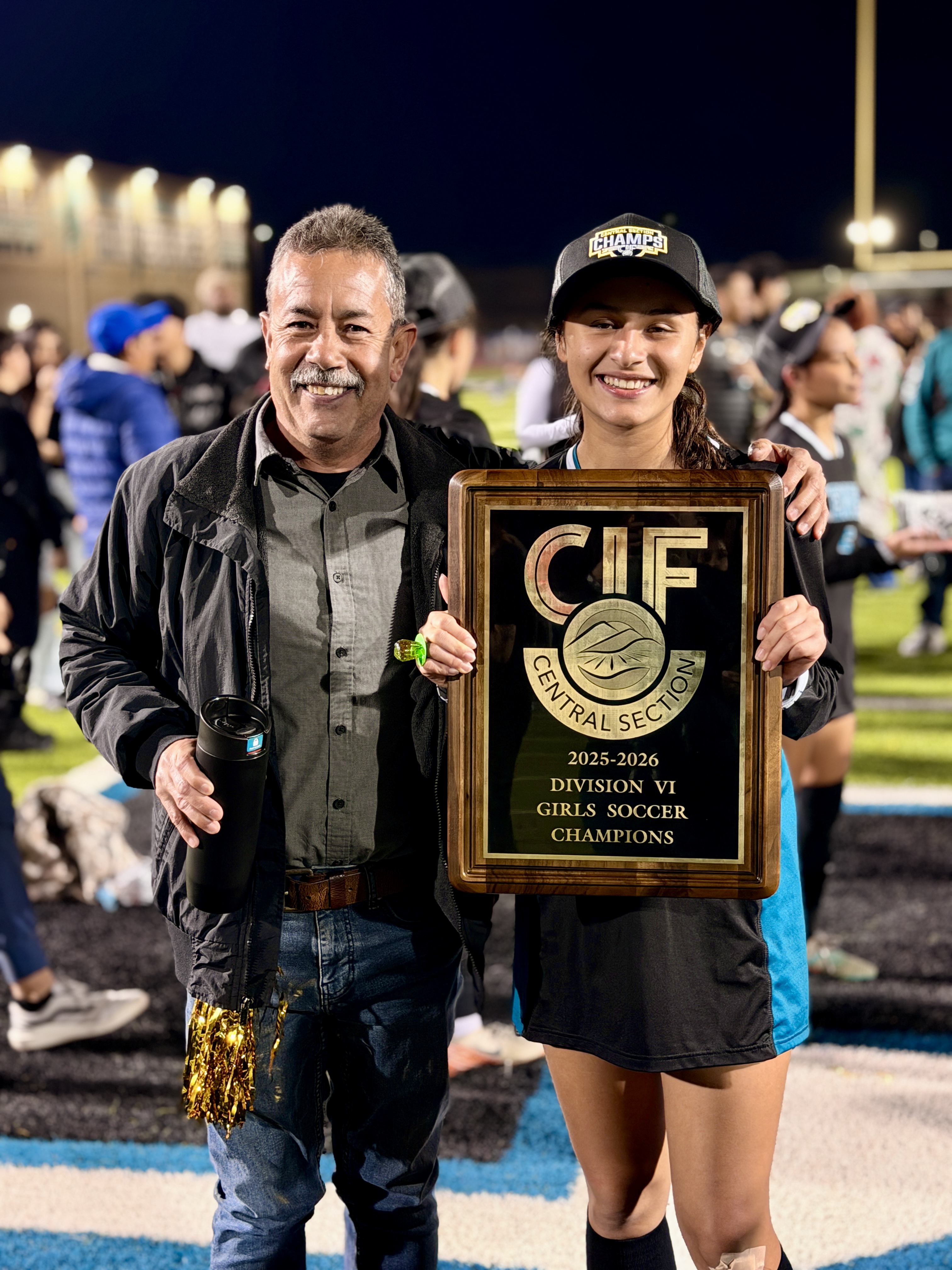 The image shows two people, a man and a woman, holding a trophy together. They appear to be celebrating the woman's achievement, as indicated by the "CIF 2023-2024 Division 2 Girls Soccer Champions" text on the trophy. The background suggests this is taking place at a sports event or field.