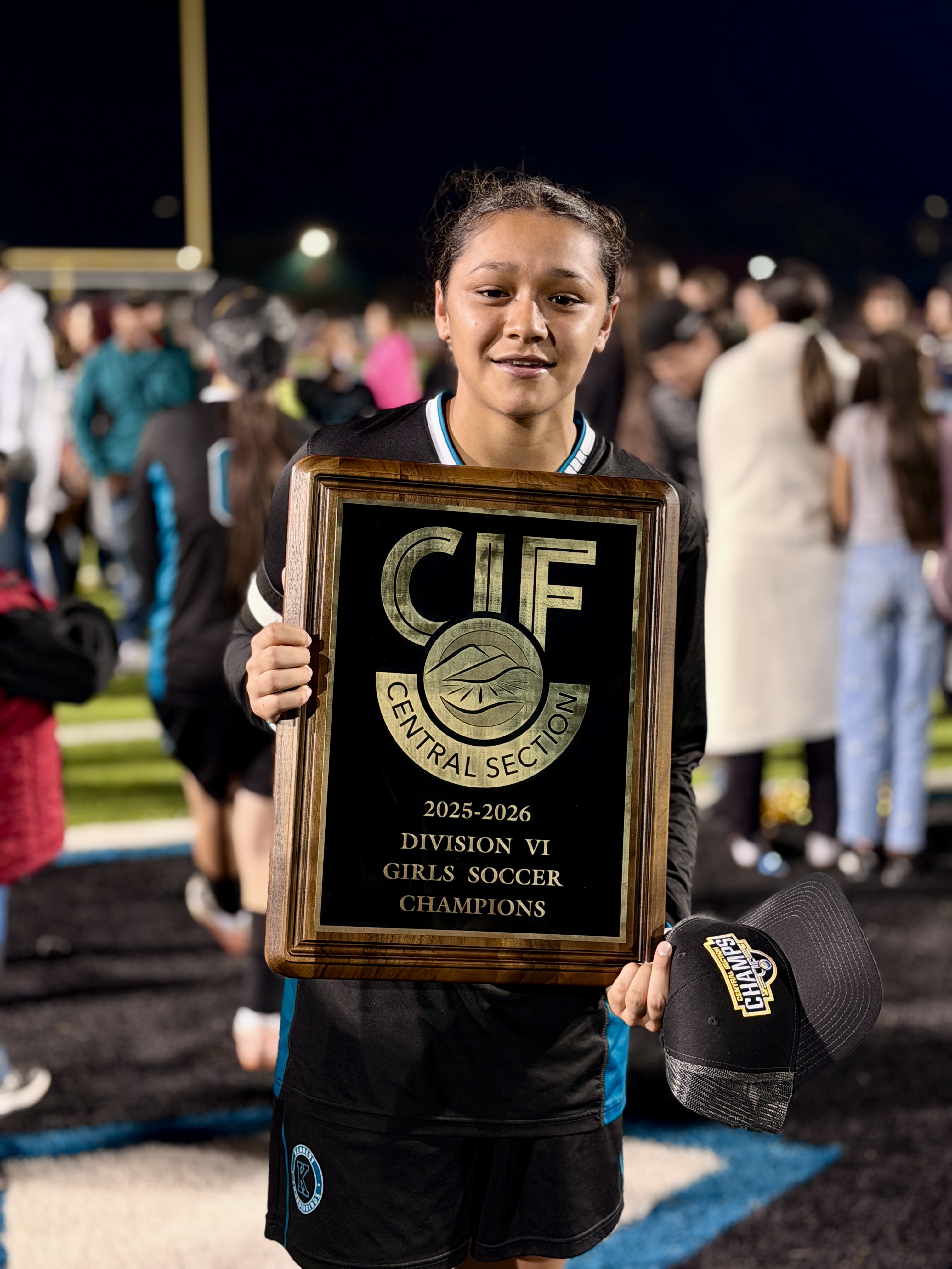 A young female athlete proudly holds a championship trophy in front of a crowd of people at what appears to be a sports event or celebration.
