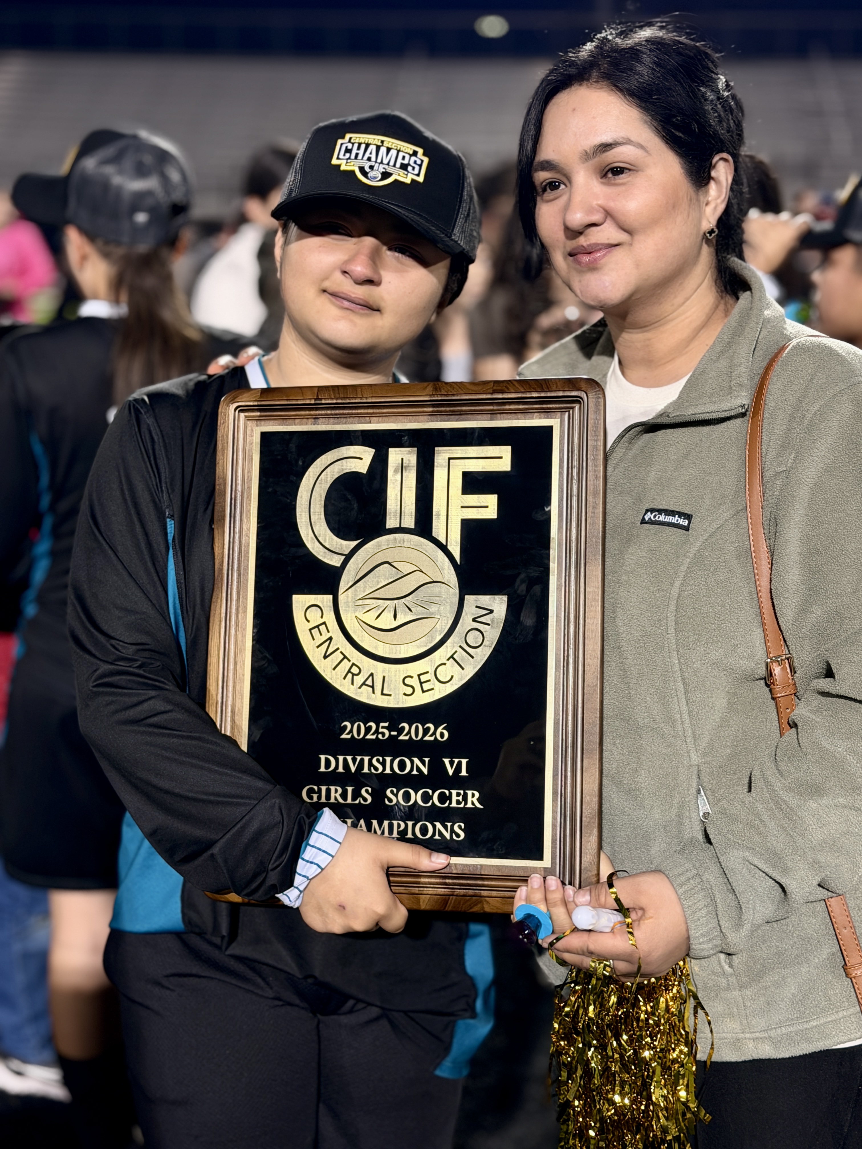 Two individuals, a young person and an adult, are holding a trophy that appears to be for a girls' soccer championship. They are both smiling and seem to be celebrating their achievement.