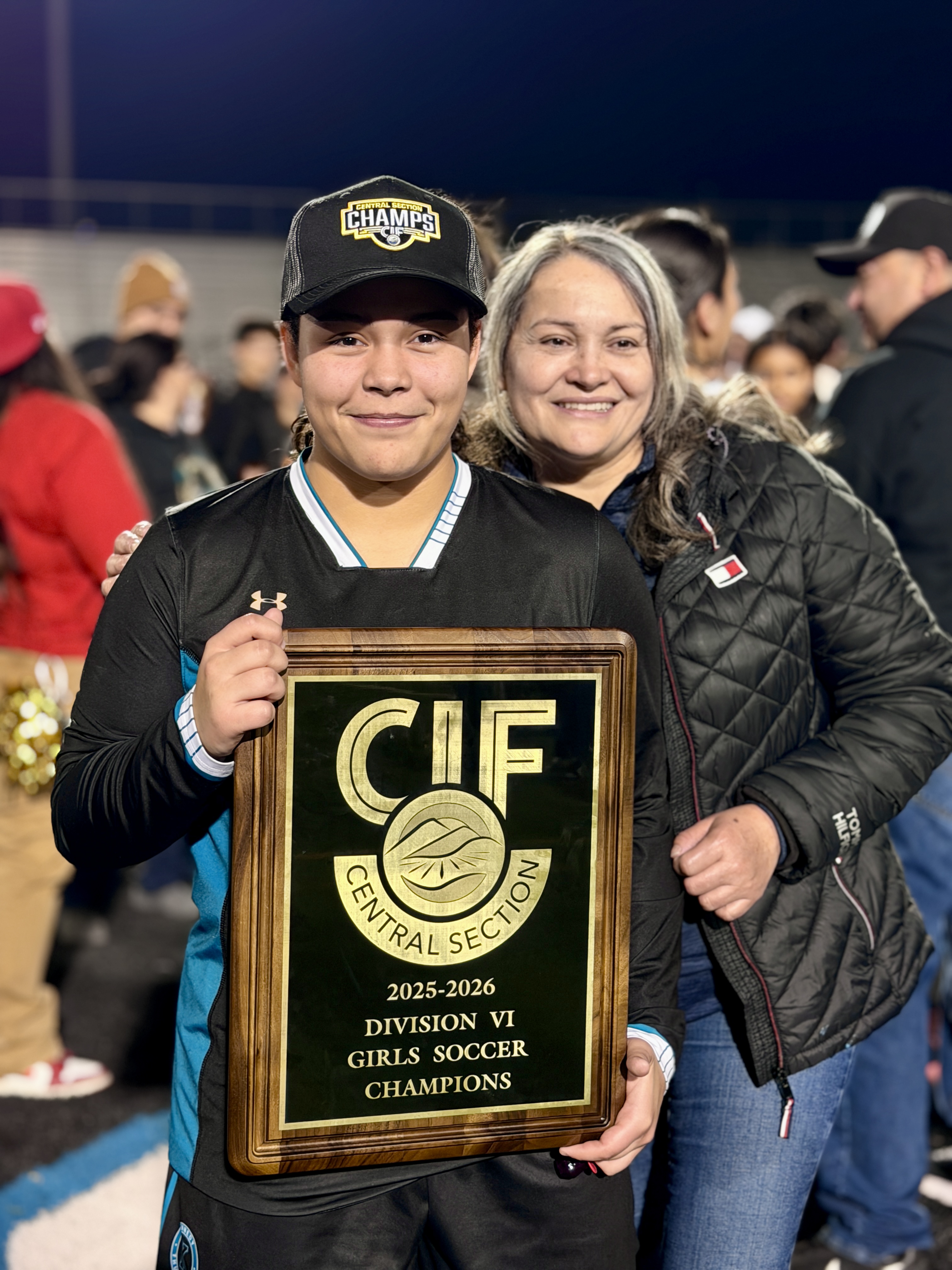 A young person holding a trophy while standing next to an adult, with a crowd of people visible in the background.