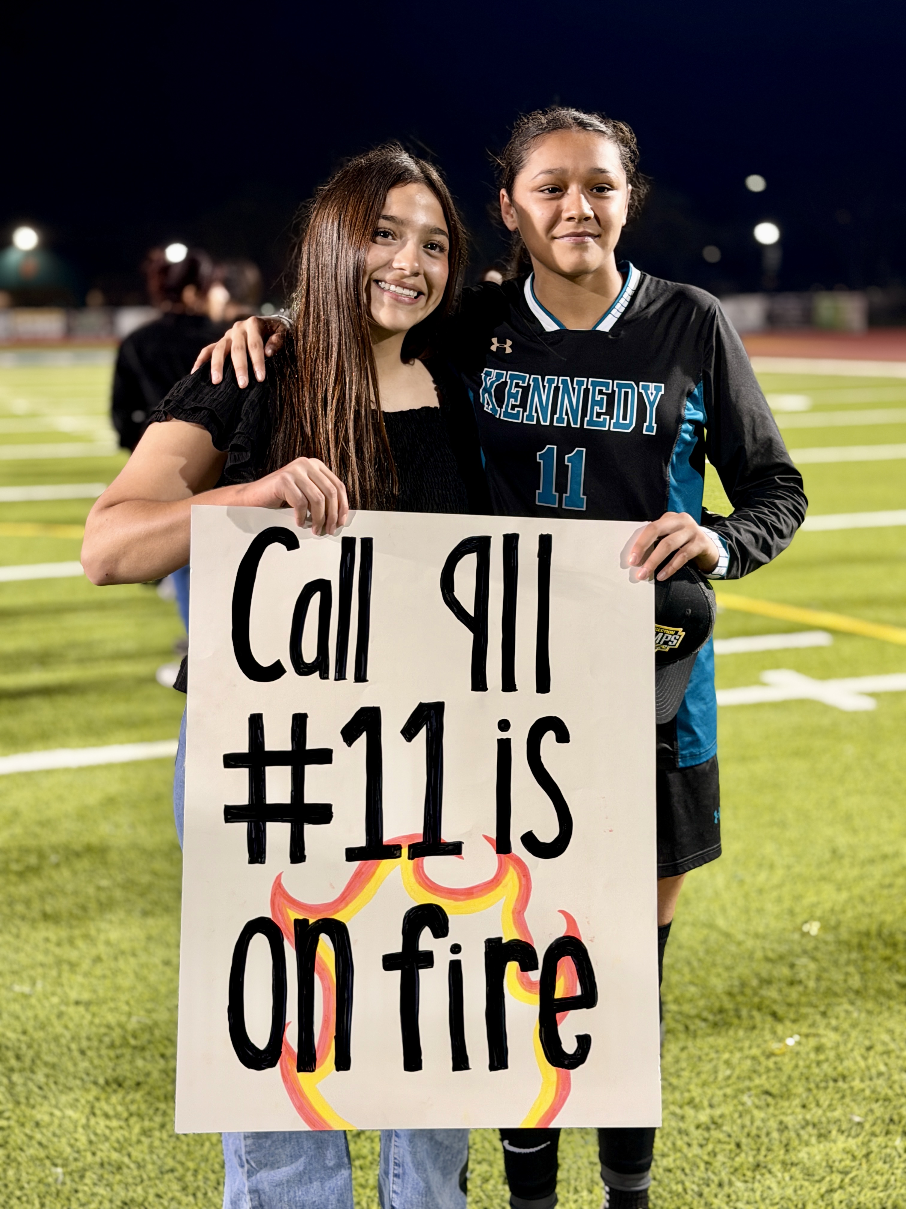 Two young women are holding a sign that says "Call all #11 is on fire" on a football field at night.