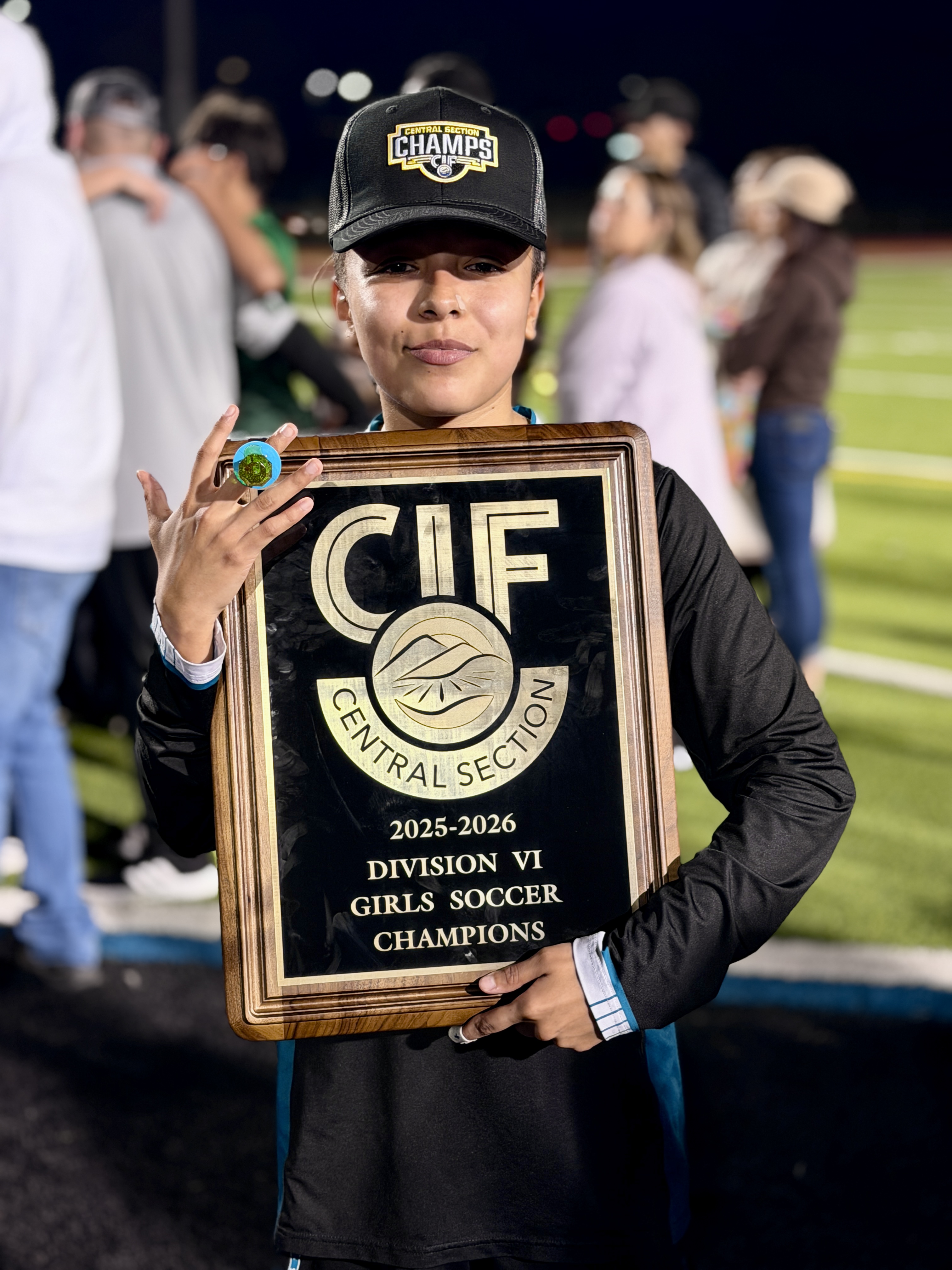A young person wearing a baseball cap and holding a trophy for the Central Illinois Federation (CIF) 2025-2026 Division VI Girls Soccer Champions.