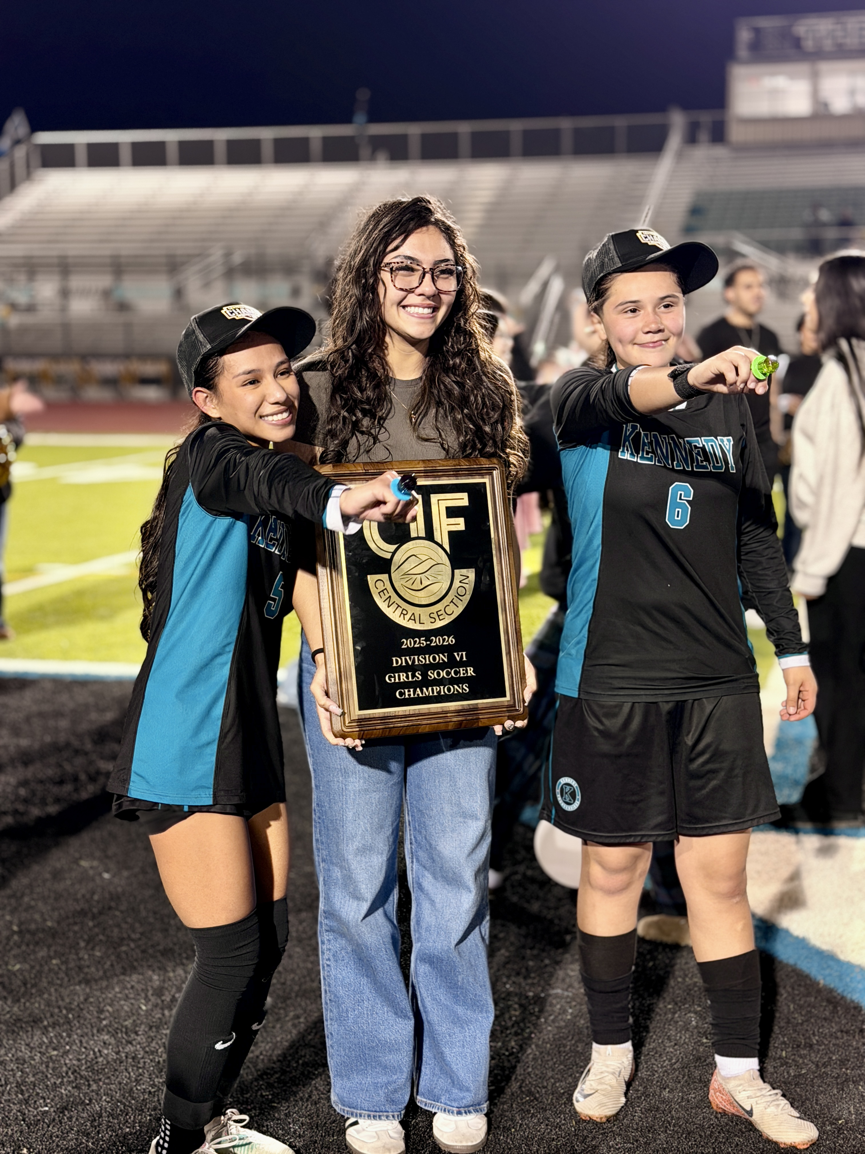 The image shows three young people, two girls and one boy, standing together and holding a trophy or award. They appear to be celebrating an accomplishment, with the background indicating they are on a sports field or stadium.