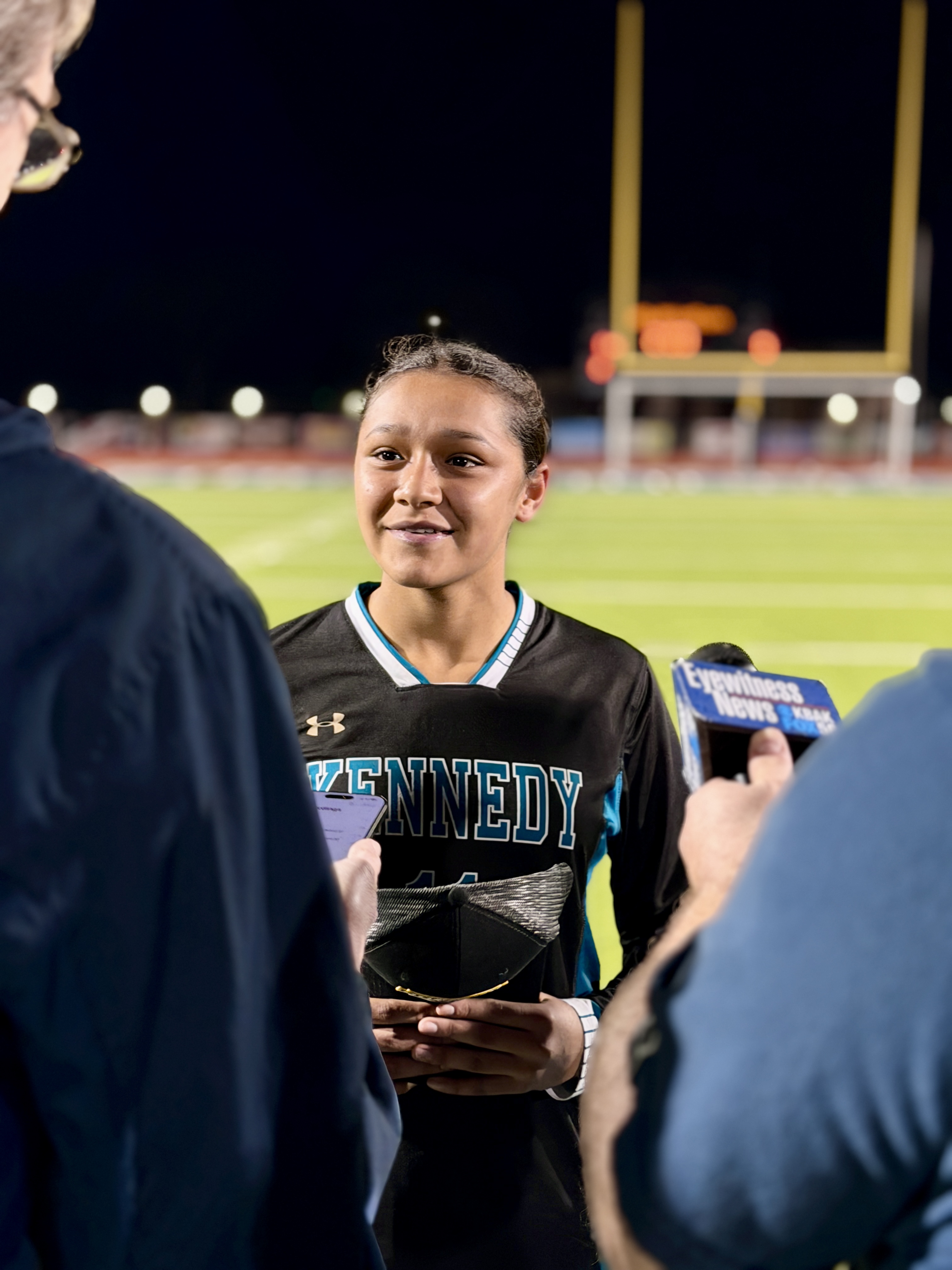 A smiling young person in a sports jersey stands on a sports field, surrounded by other people.