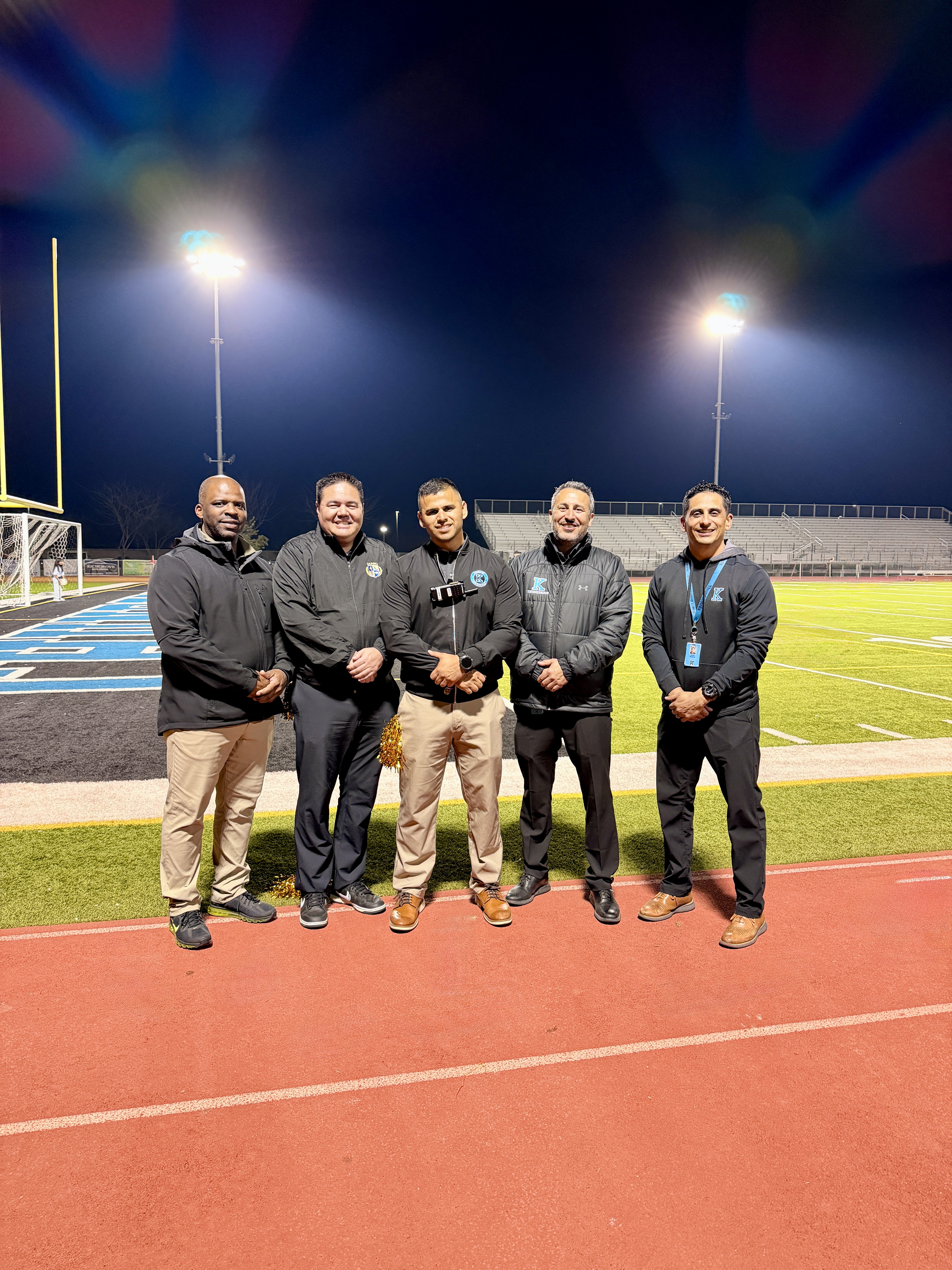 A group of five men in casual attire standing on a brightly lit sports field at night, with stadium lights illuminating the scene.