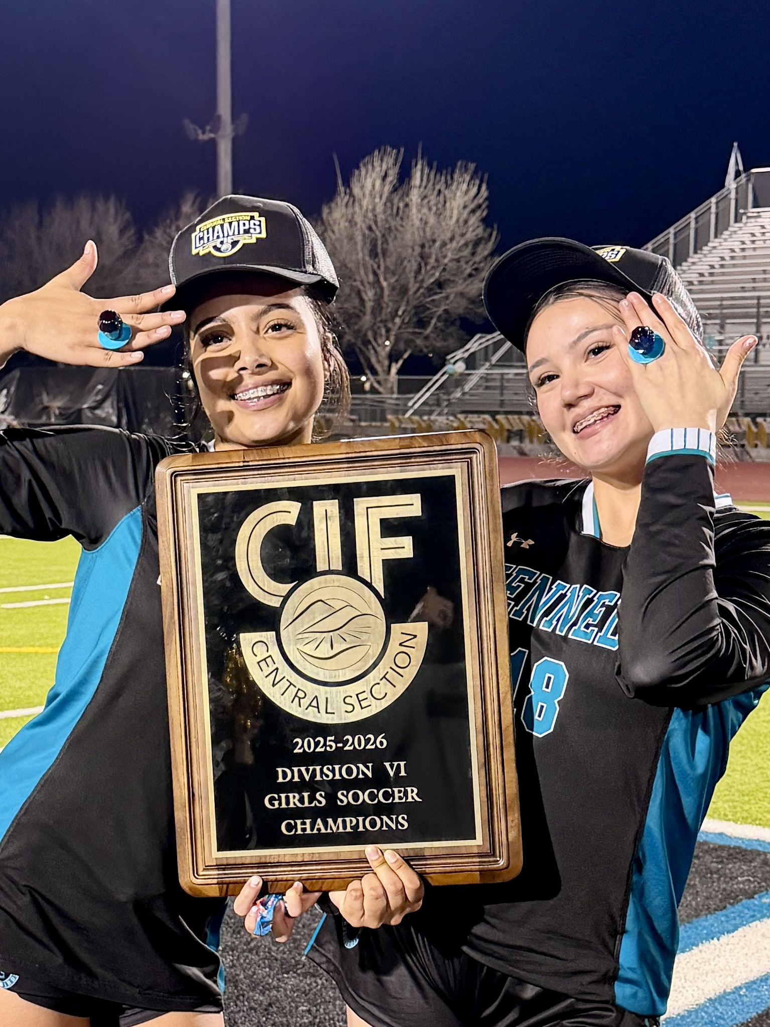 Two smiling young women holding a framed award for Division VI girls soccer champions on a sports field at night.