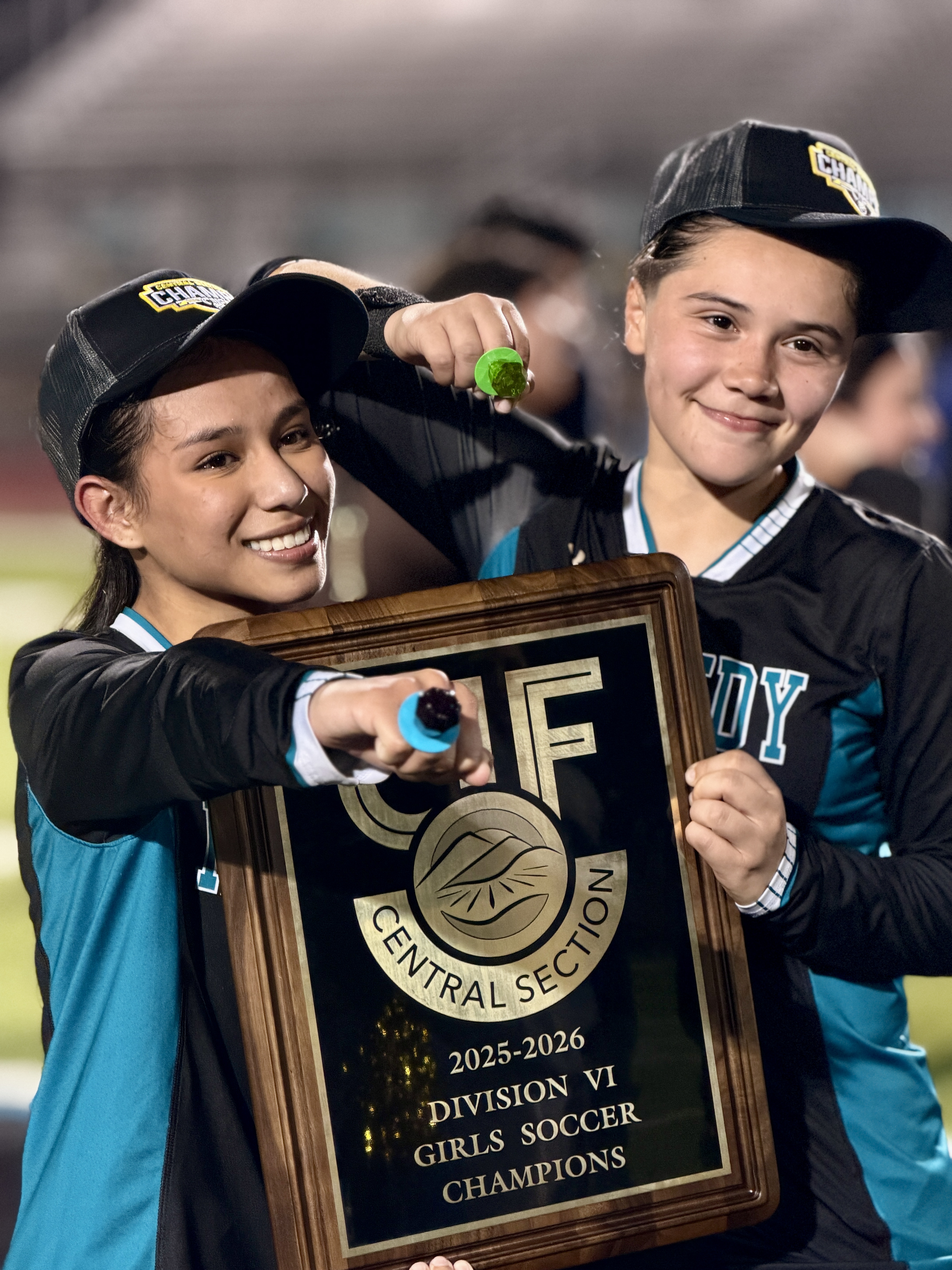 Two young girls in sports uniforms are holding a trophy and celebrating their victory as girls soccer champions for the 2025-2026 division.