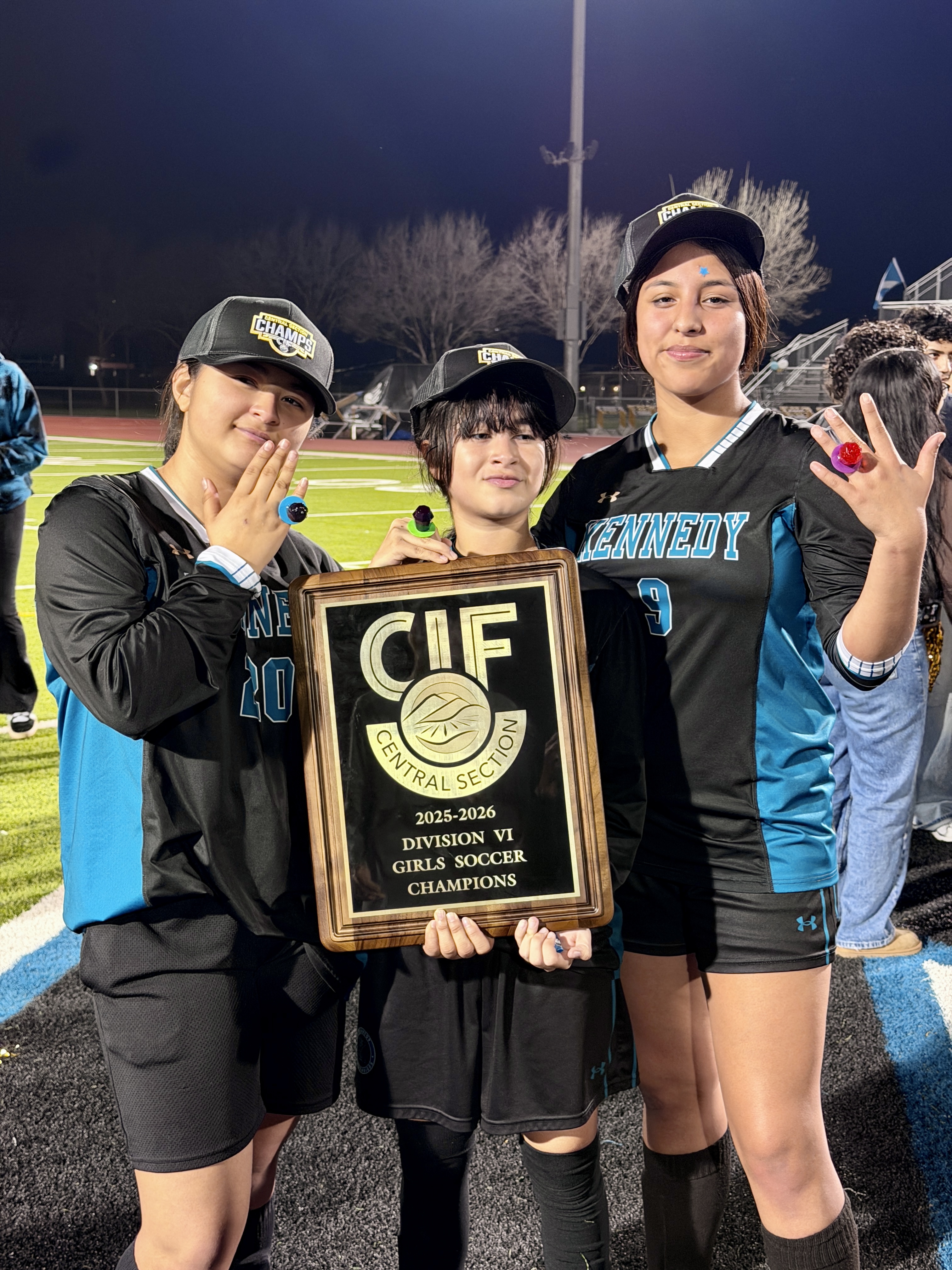 Three young women in sports uniforms are holding a trophy and celebrating their victory on a sports field at night.