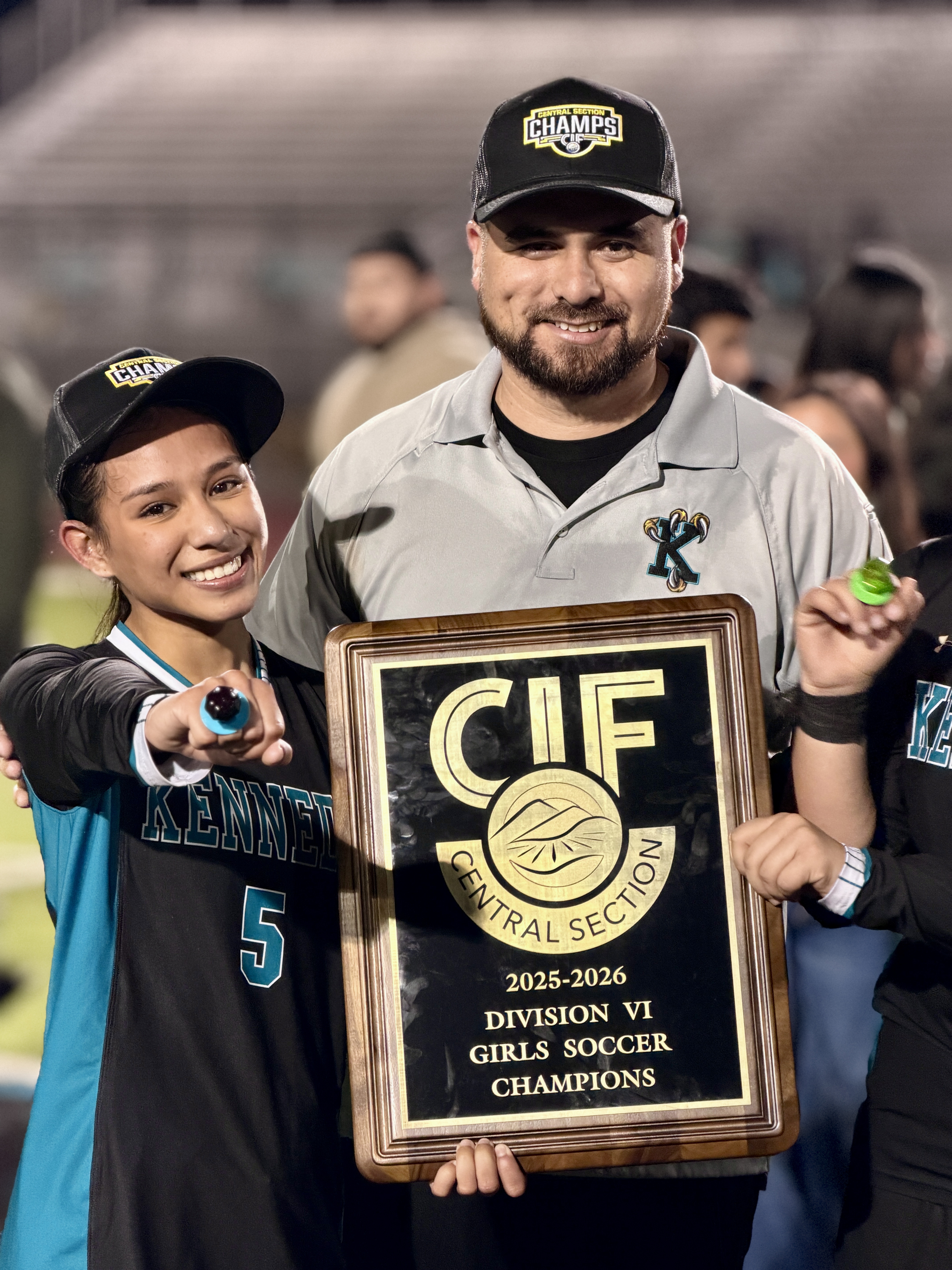 The image shows two individuals, a man and a young boy, holding a trophy and celebrating their victory as the 2025-2026 Division VI Girls Soccer Champions.