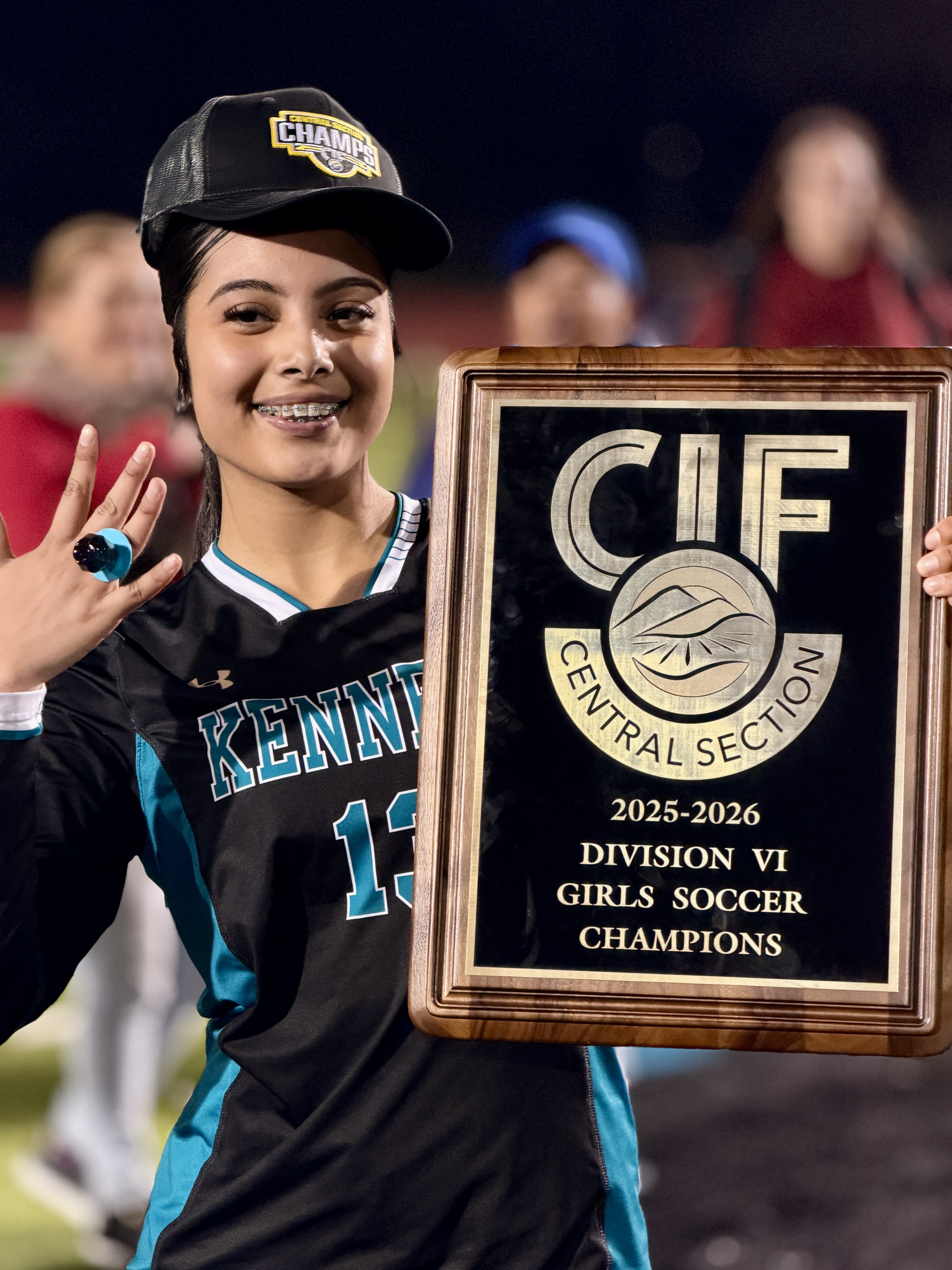 A young girl in a sports jersey and cap is holding a trophy and smiling, with a crowd of people visible in the background.