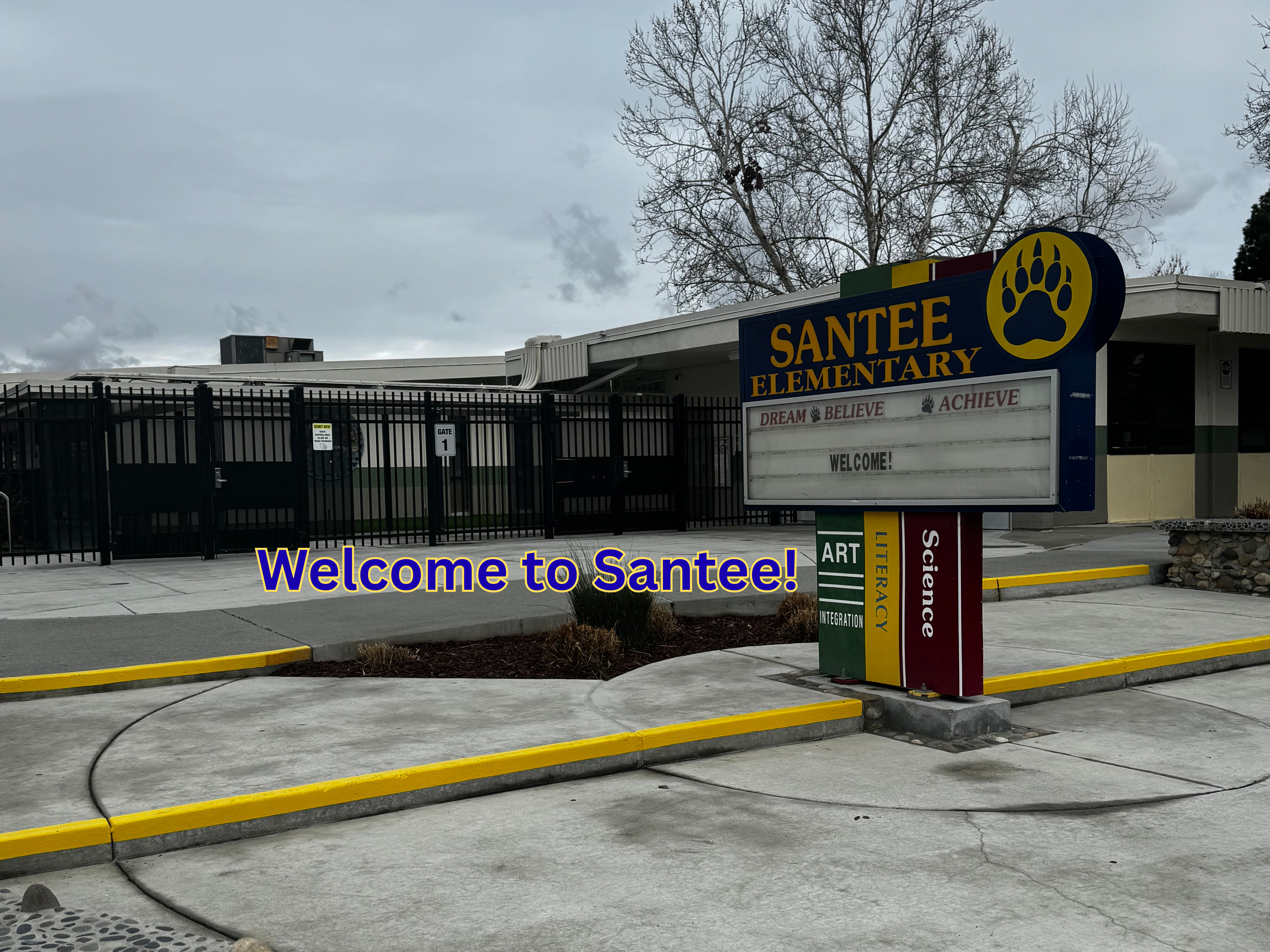 A welcome sign for Santee Elementary School stands in the foreground with the school building and a fence in the background.