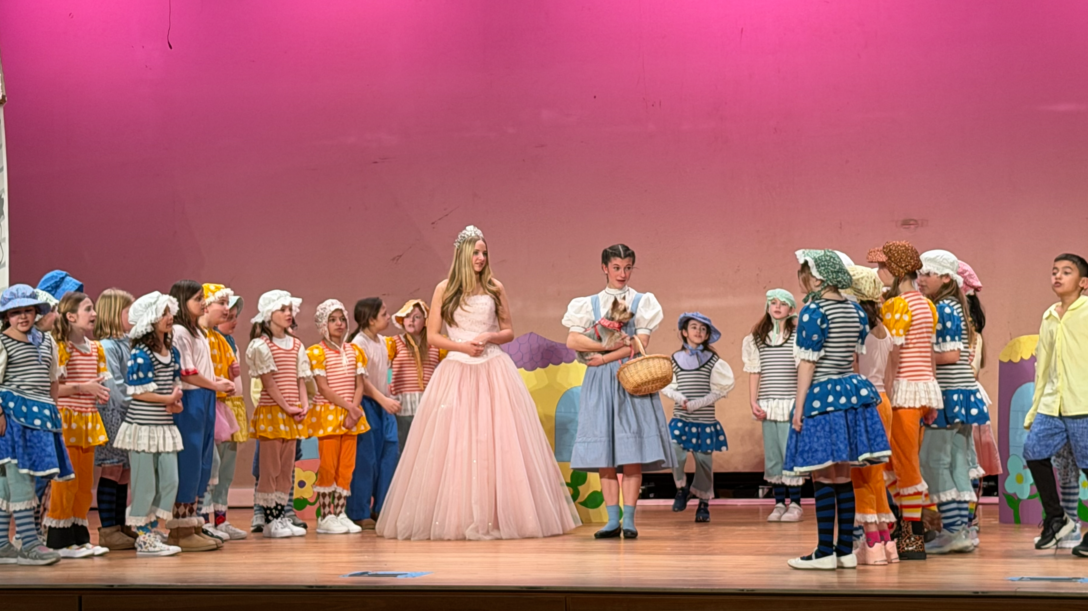 A group of colorfully dressed performers, including a woman in a pink dress, stand on a stage with a vibrant pink backdrop.