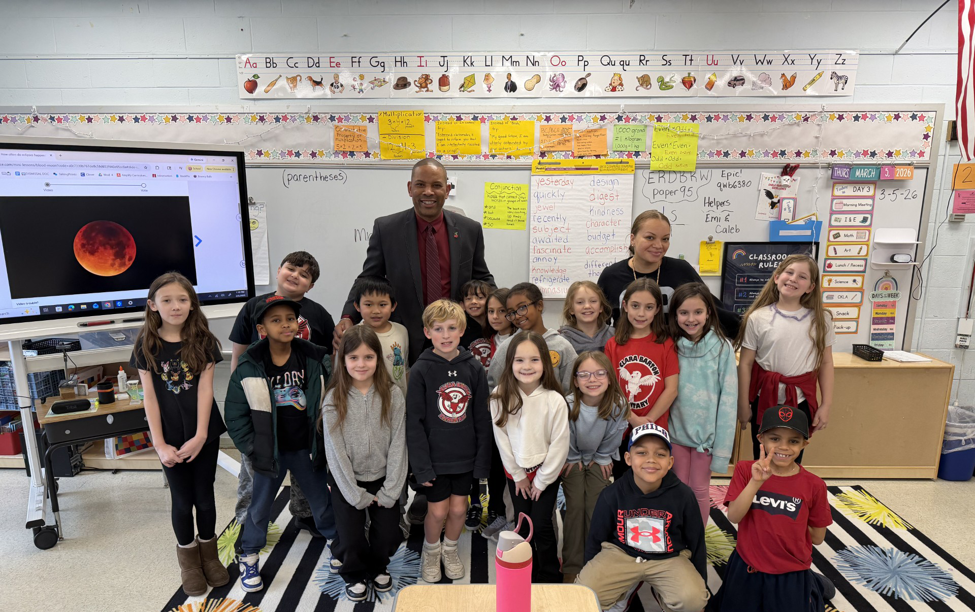 The image shows a group of students and a teacher standing in a classroom setting, with various educational materials and displays visible in the background.