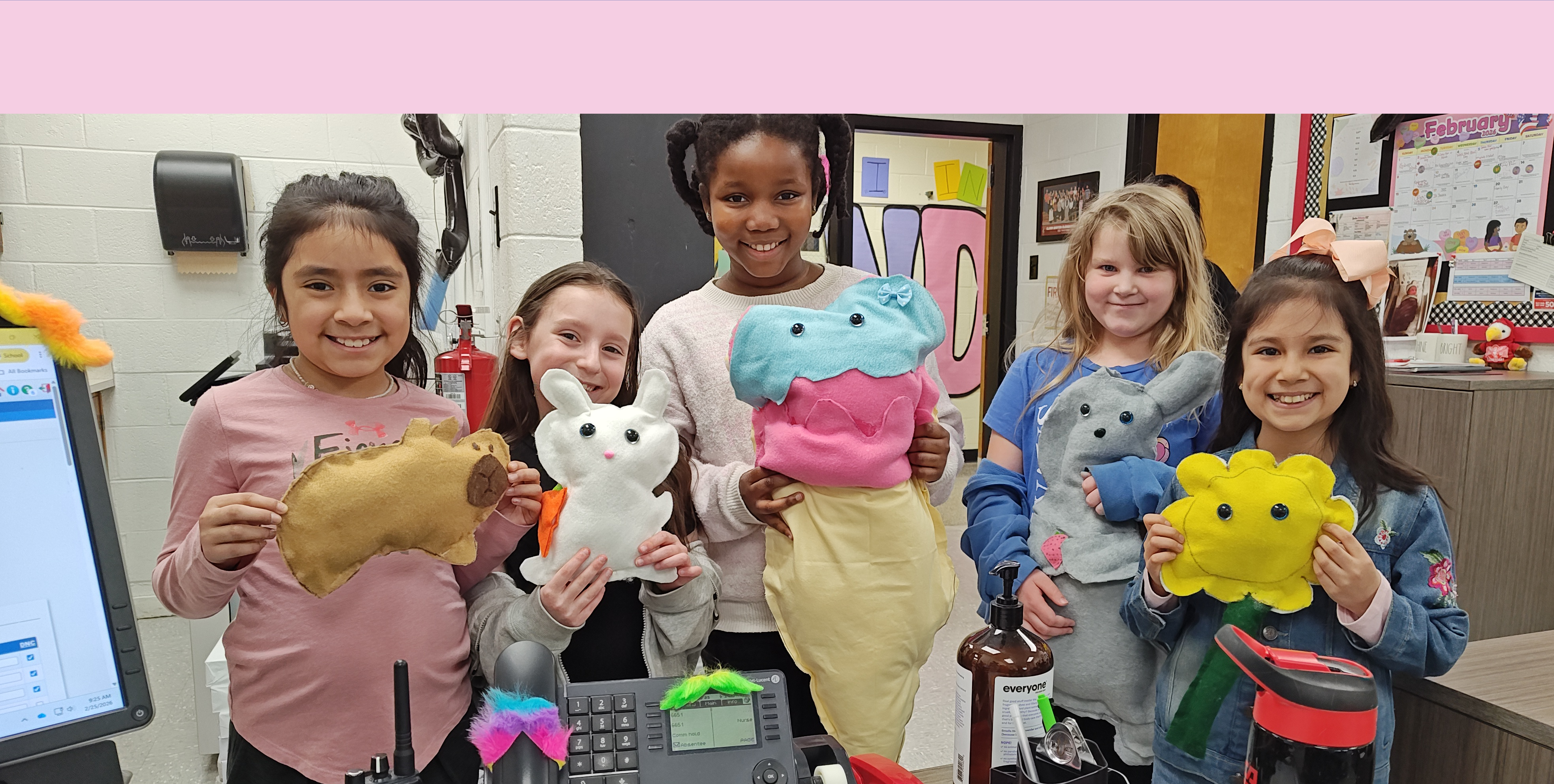 A group of smiling children, each holding a colorful stuffed animal, stand together in what appears to be a classroom setting with various educational materials visible in the background.