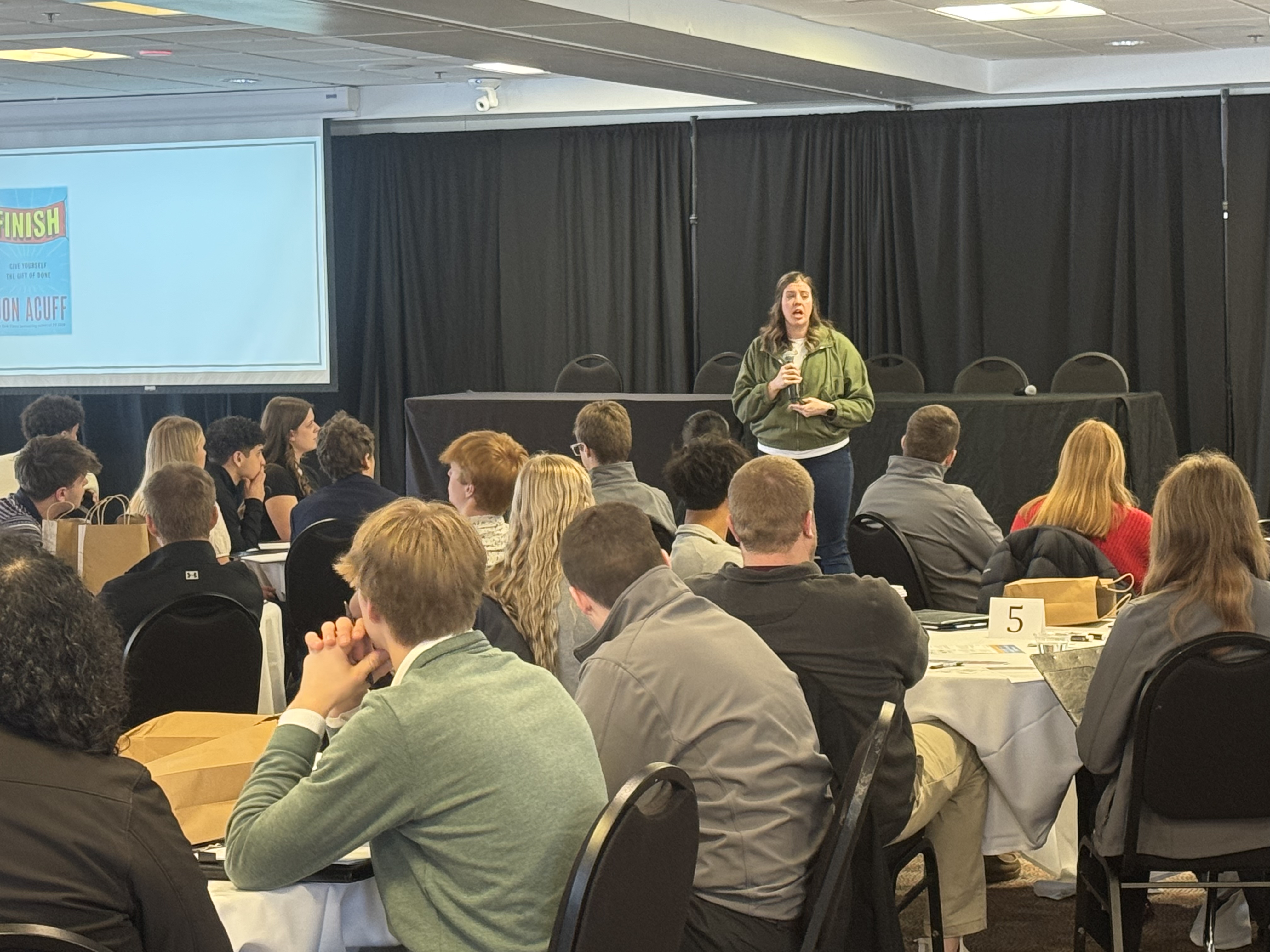 A woman is standing at a podium in front of a large audience seated at tables in a conference room setting.