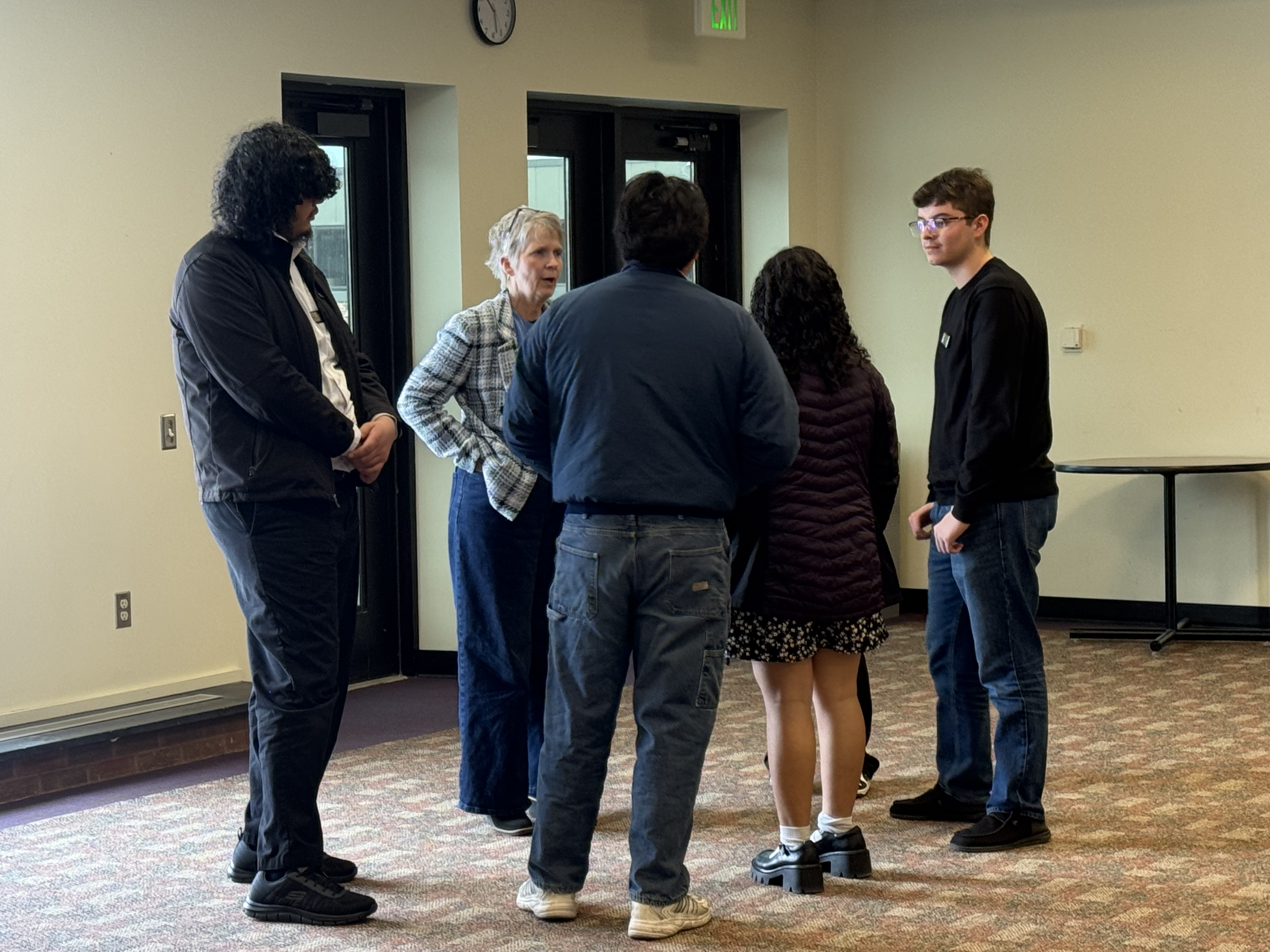A group of people, including a woman with short gray hair, are gathered in what appears to be a room or hallway, engaged in conversation.
