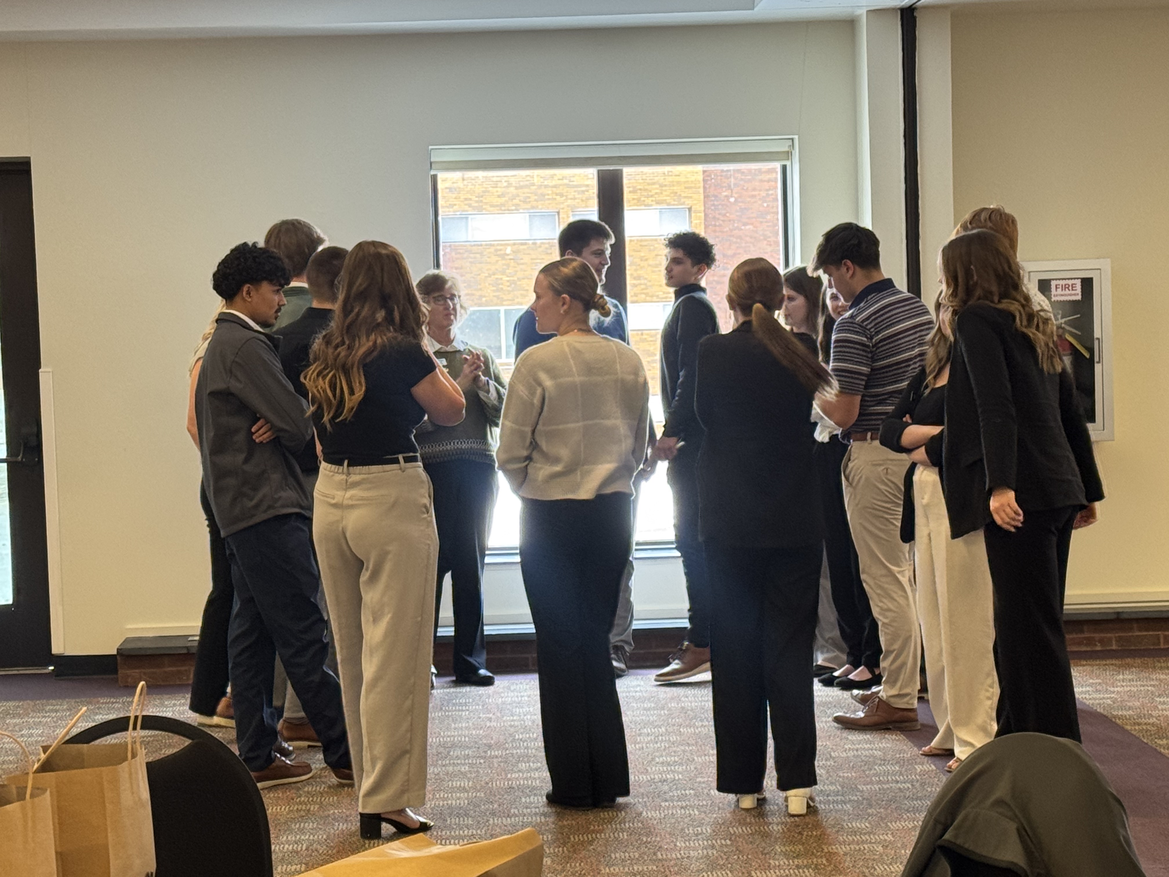 A group of people, mostly young adults, are gathered in a room with a window in the background, engaged in conversation.