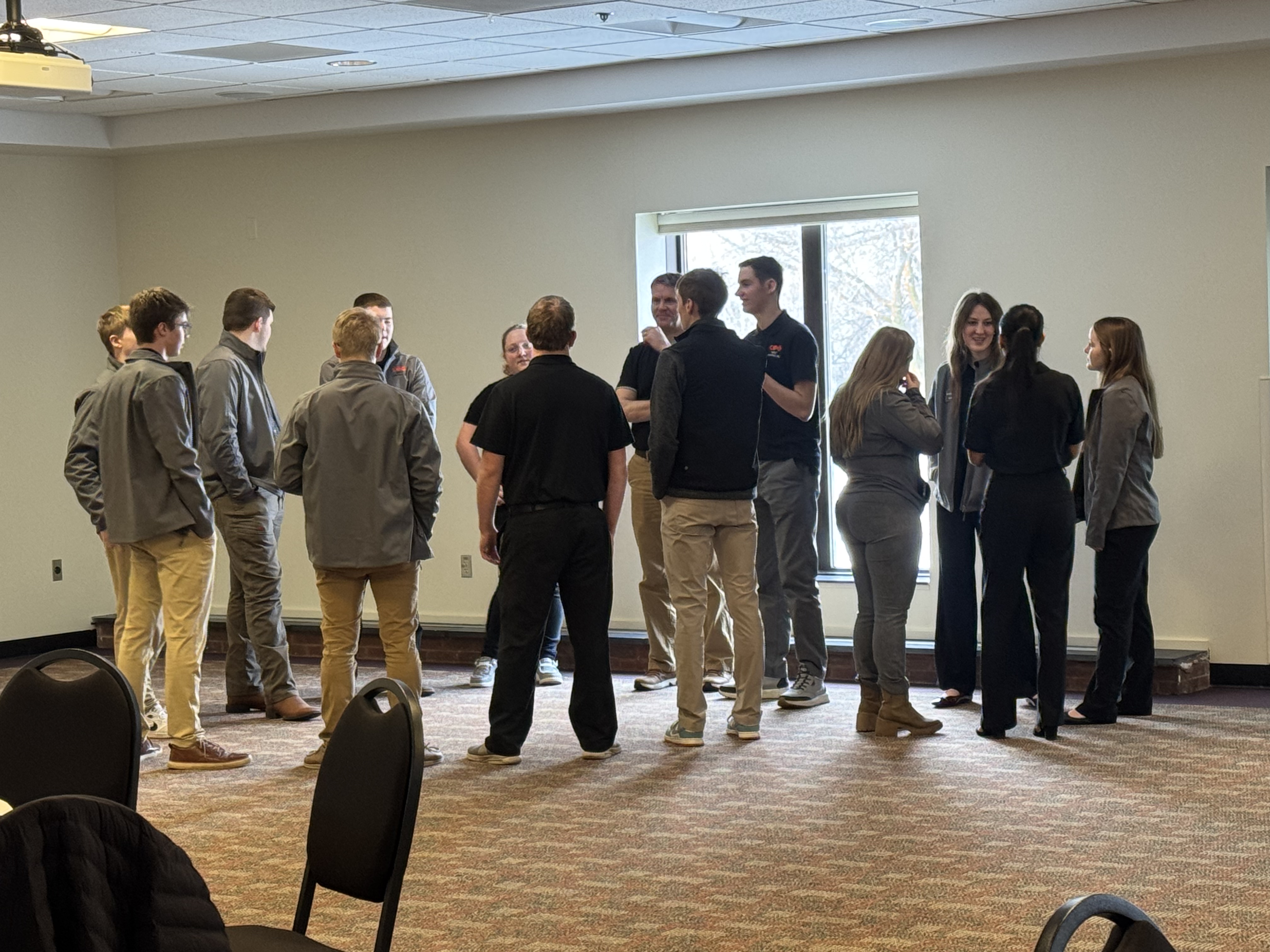 A group of people, mostly young adults, are gathered in a room with a tiled floor and a window visible in the background.