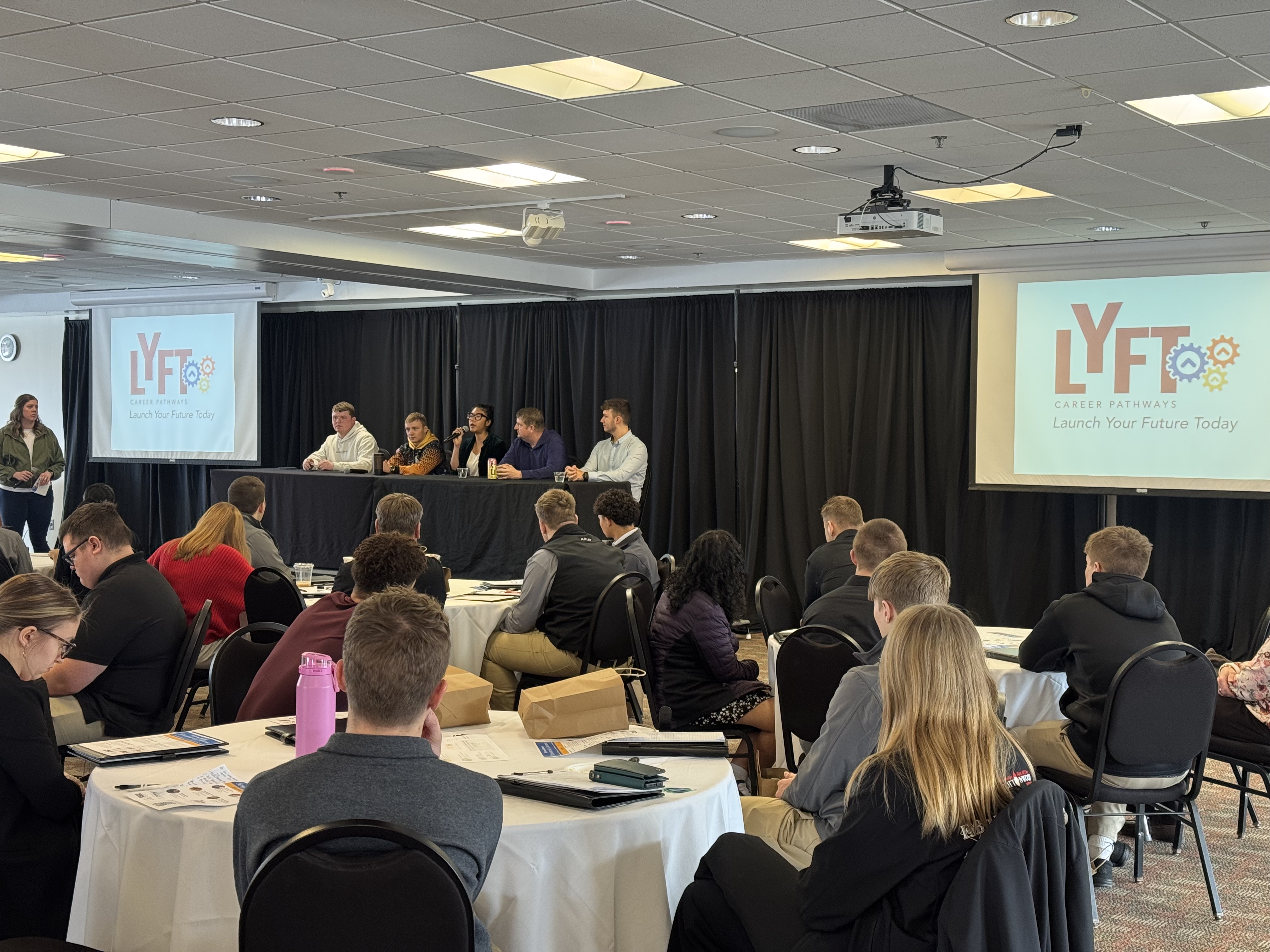 A group of people seated at tables in a conference room, with a panel of speakers on a stage at the front and a large projection screen displaying the event's logo.