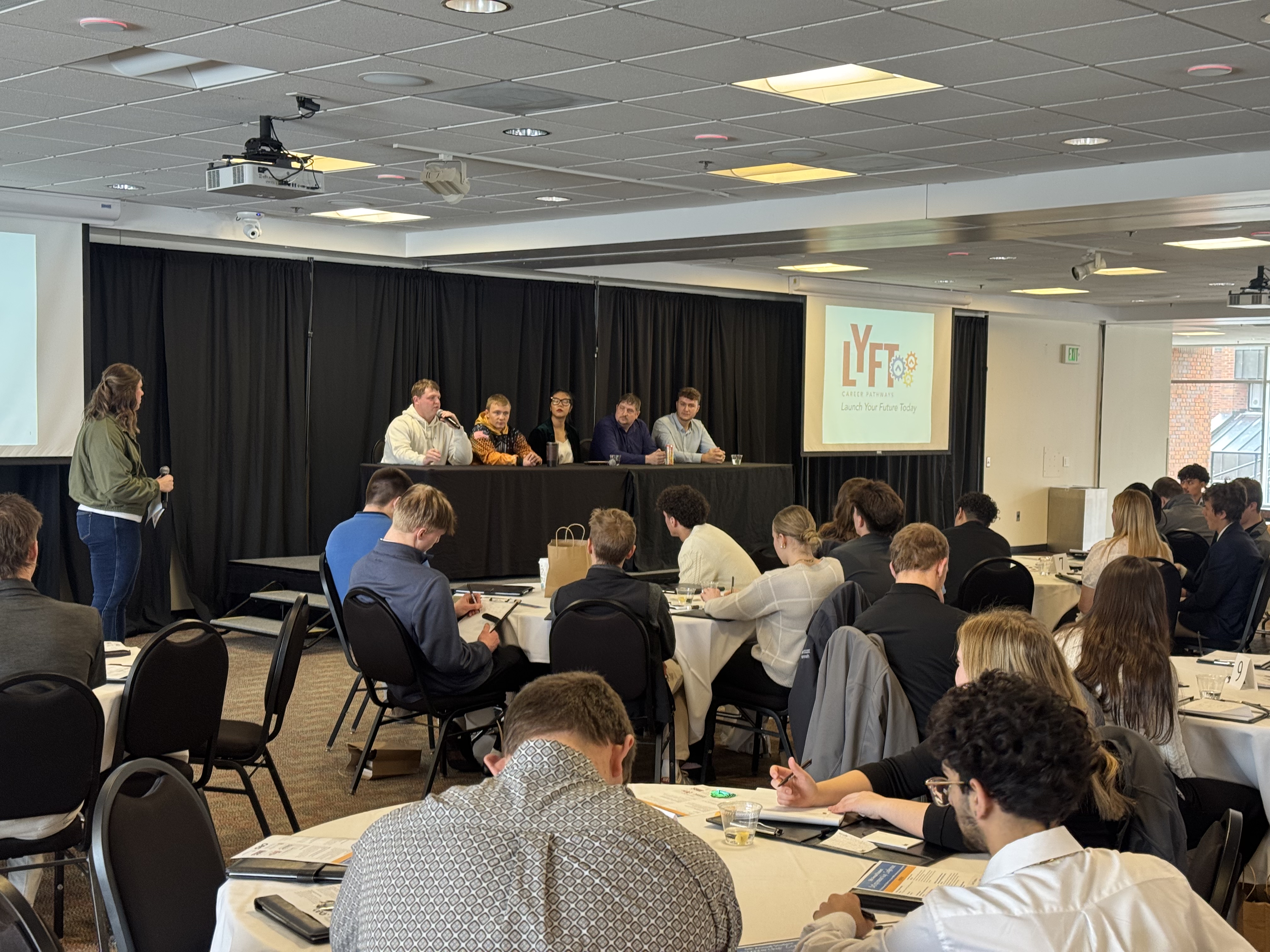 A group of people seated at tables in a conference room, with a panel of speakers on a stage in the background.