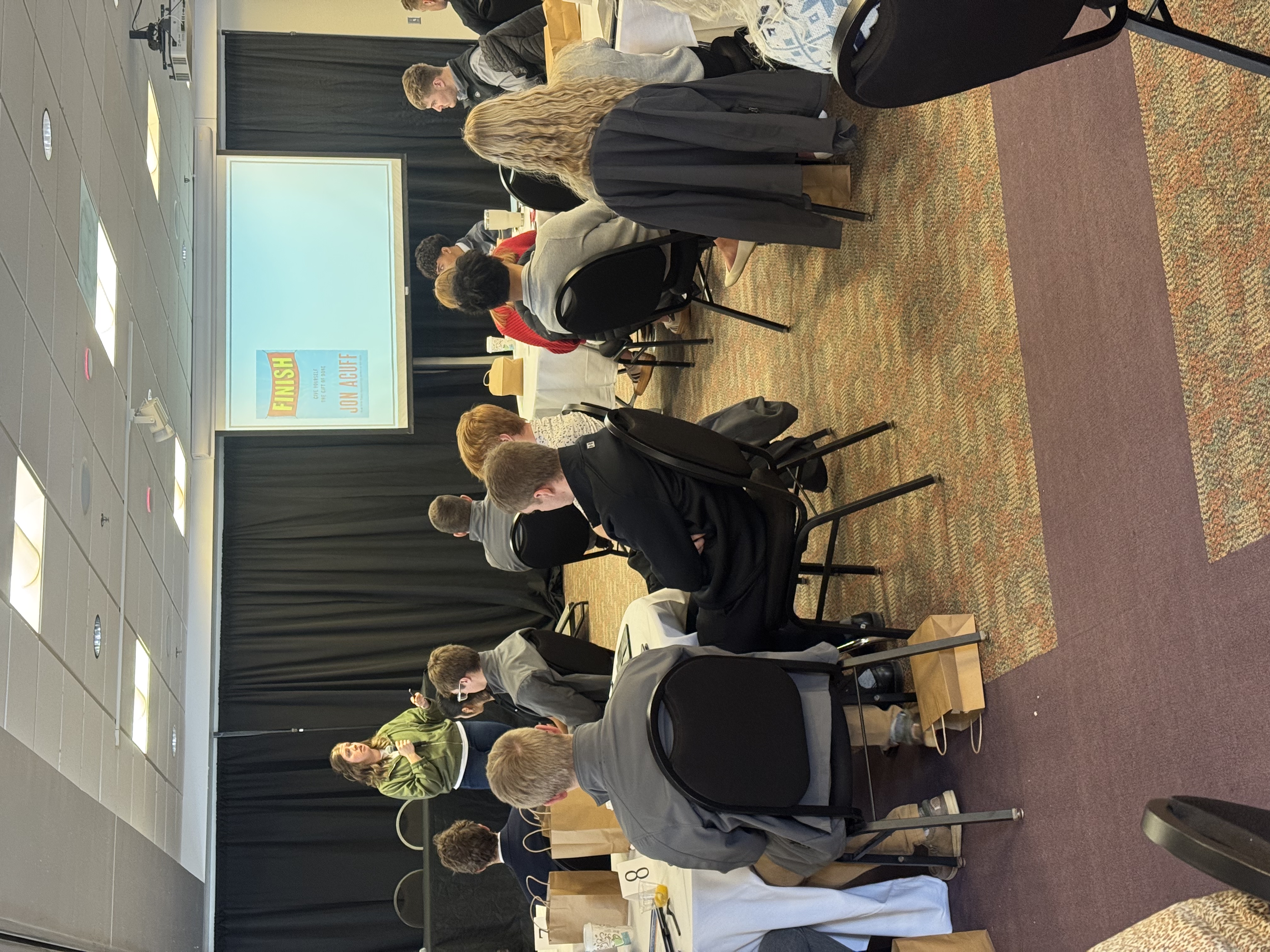 A group of people seated at tables, listening to a presentation displayed on a screen in the background.