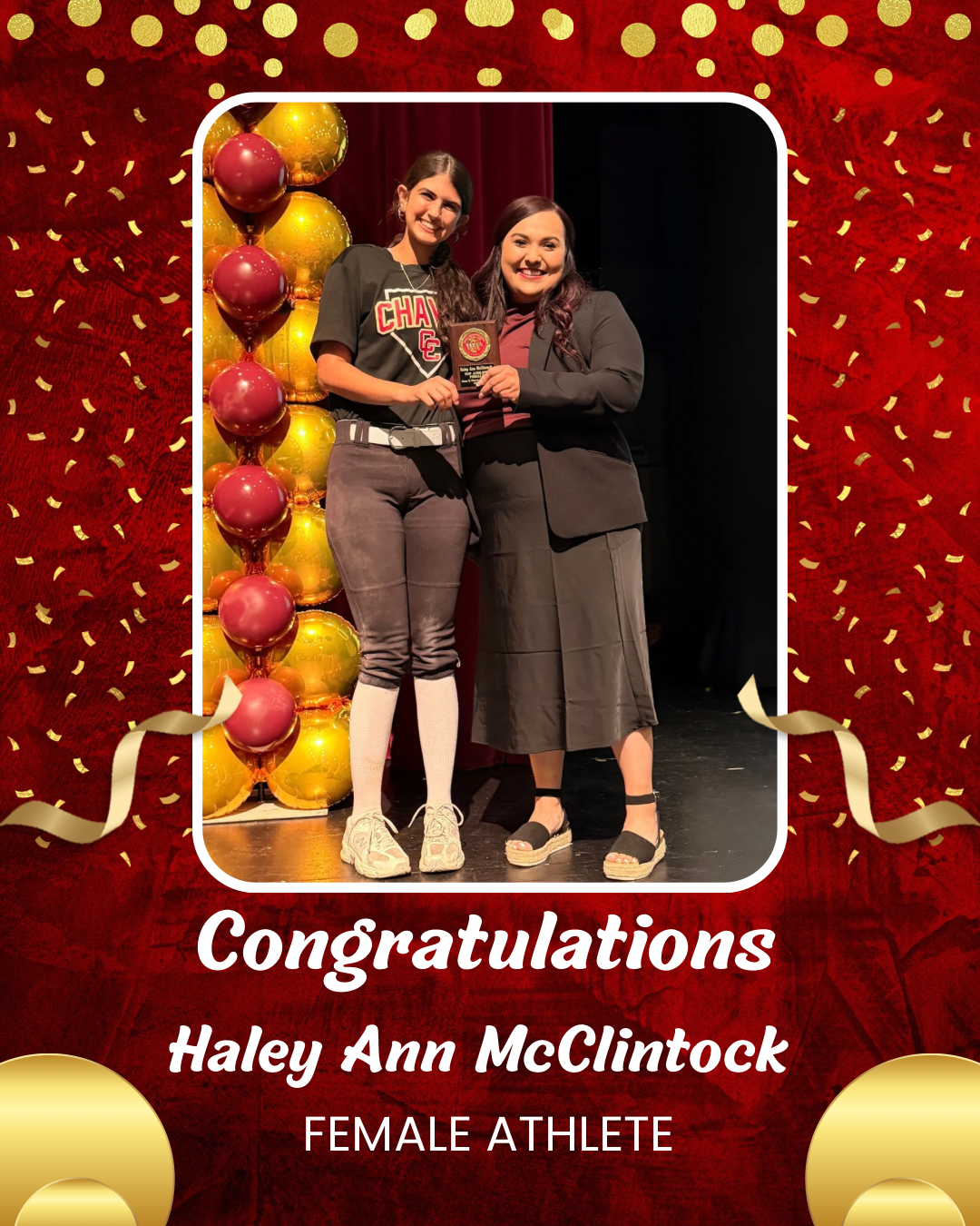 The image shows two women, one younger and one older, standing together and smiling while holding a trophy. The background is a red and yellow confetti-filled stage, with balloons in the shape of apples or other fruit.