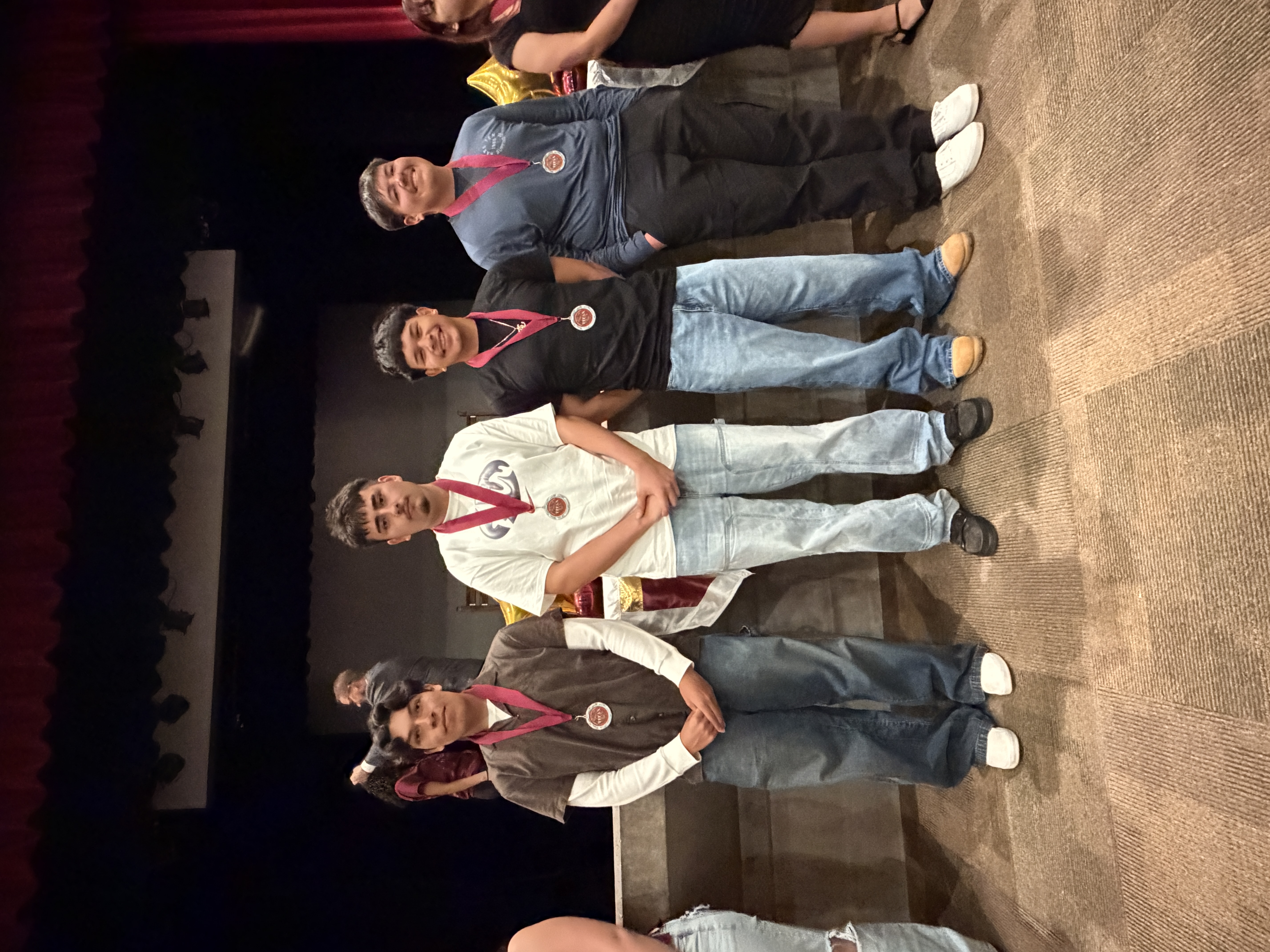 The image shows four individuals standing on a stage, wearing medals and casual clothing, with a curtain and stage backdrop visible in the background.