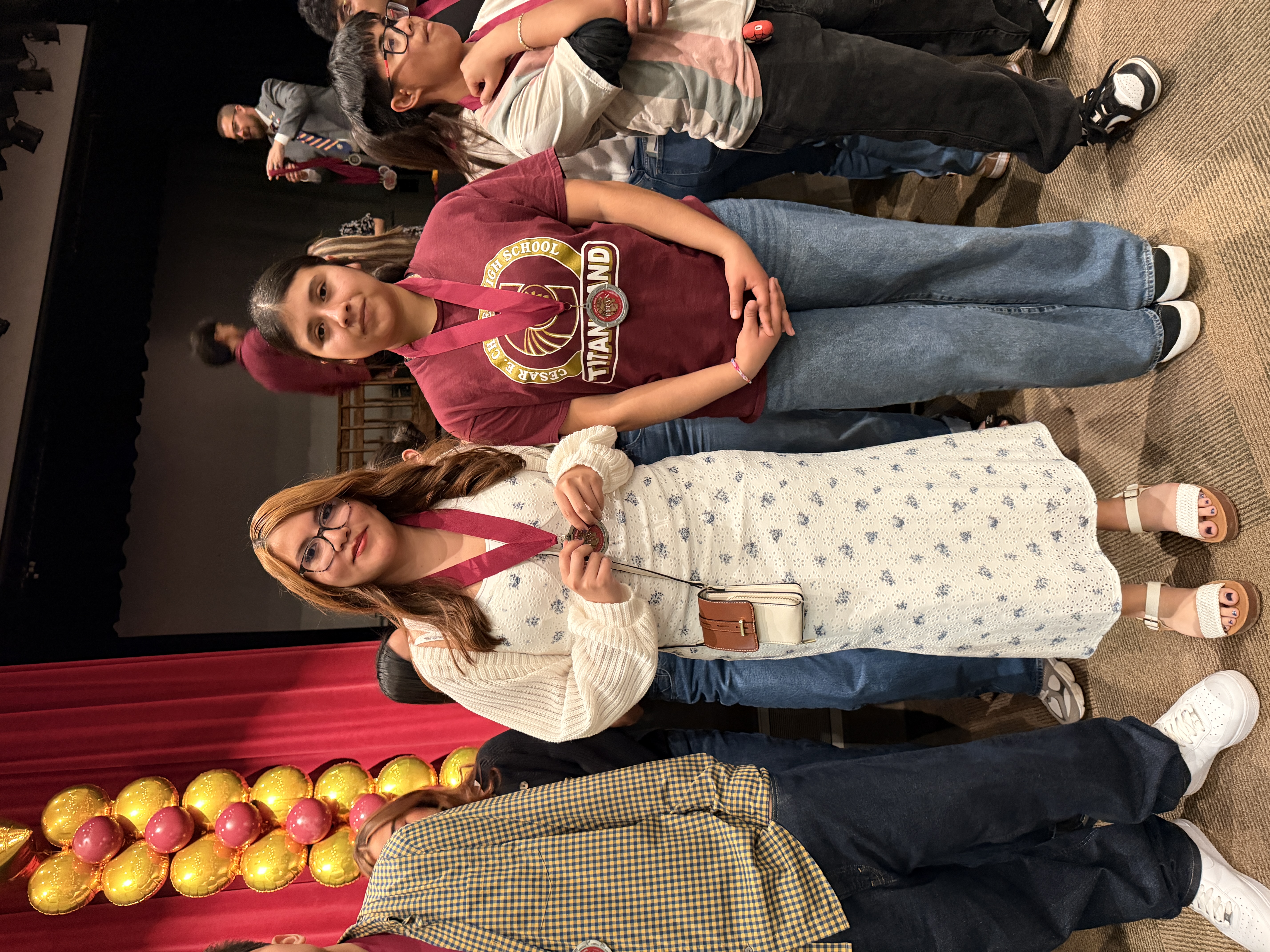 Two young women, one wearing a white sweater and the other a red shirt, stand together in front of a red curtain backdrop, with other people visible in the background.