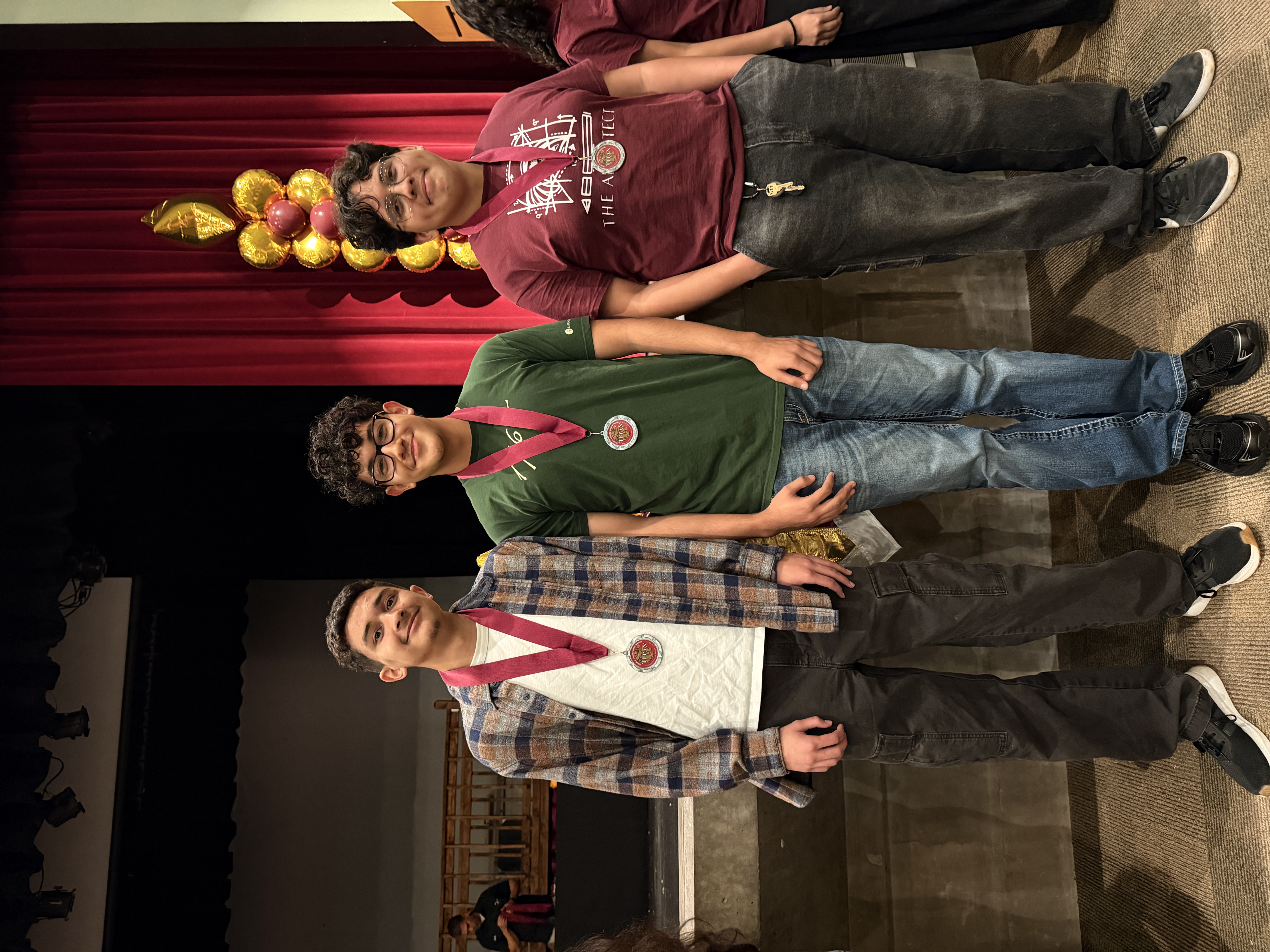 Three young men standing in front of a red curtain backdrop, with one wearing a medal around his neck.