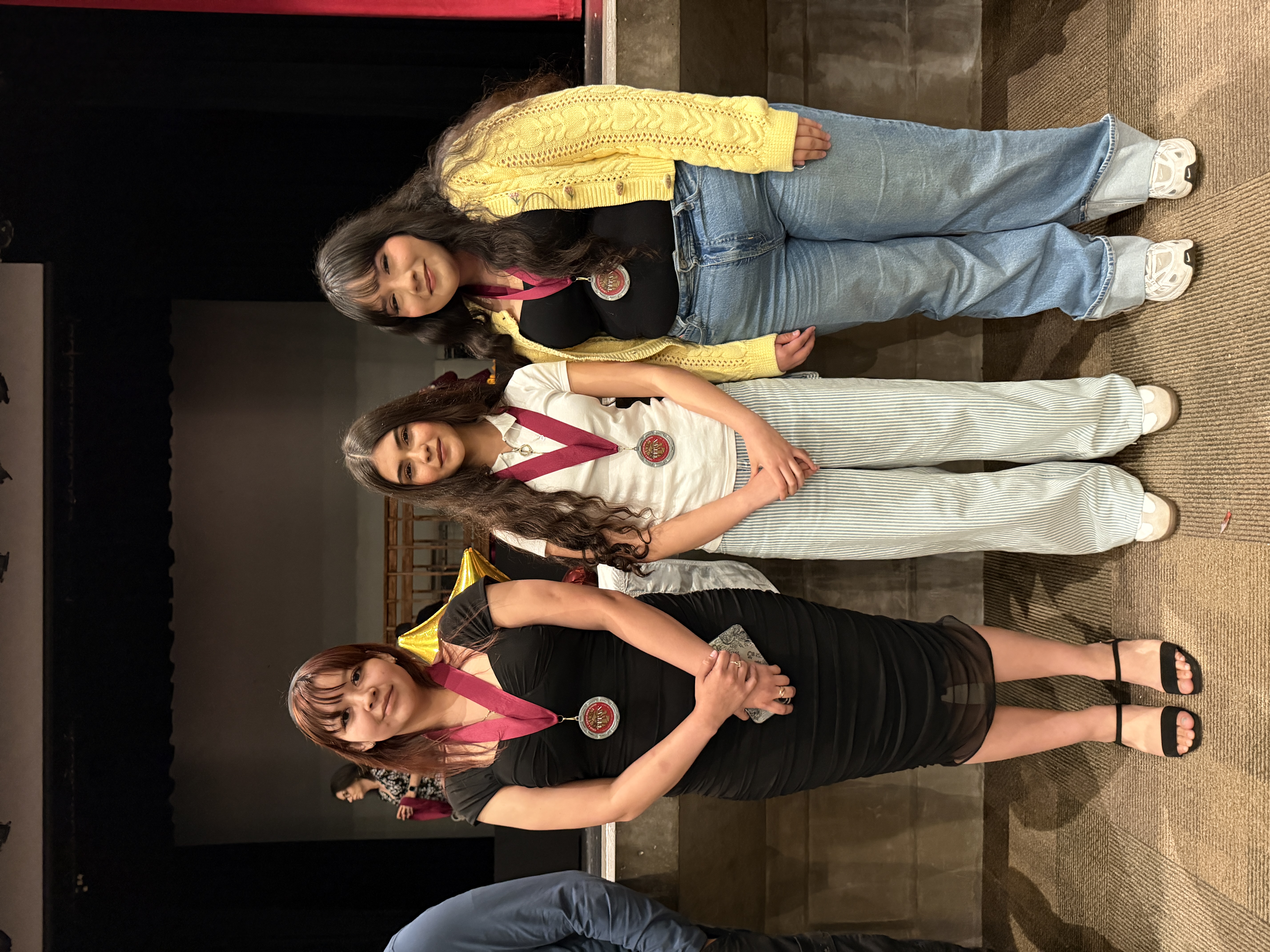 Three young women, each wearing different outfits, stand together in what appears to be a stage or performance setting, with a backdrop visible behind them.