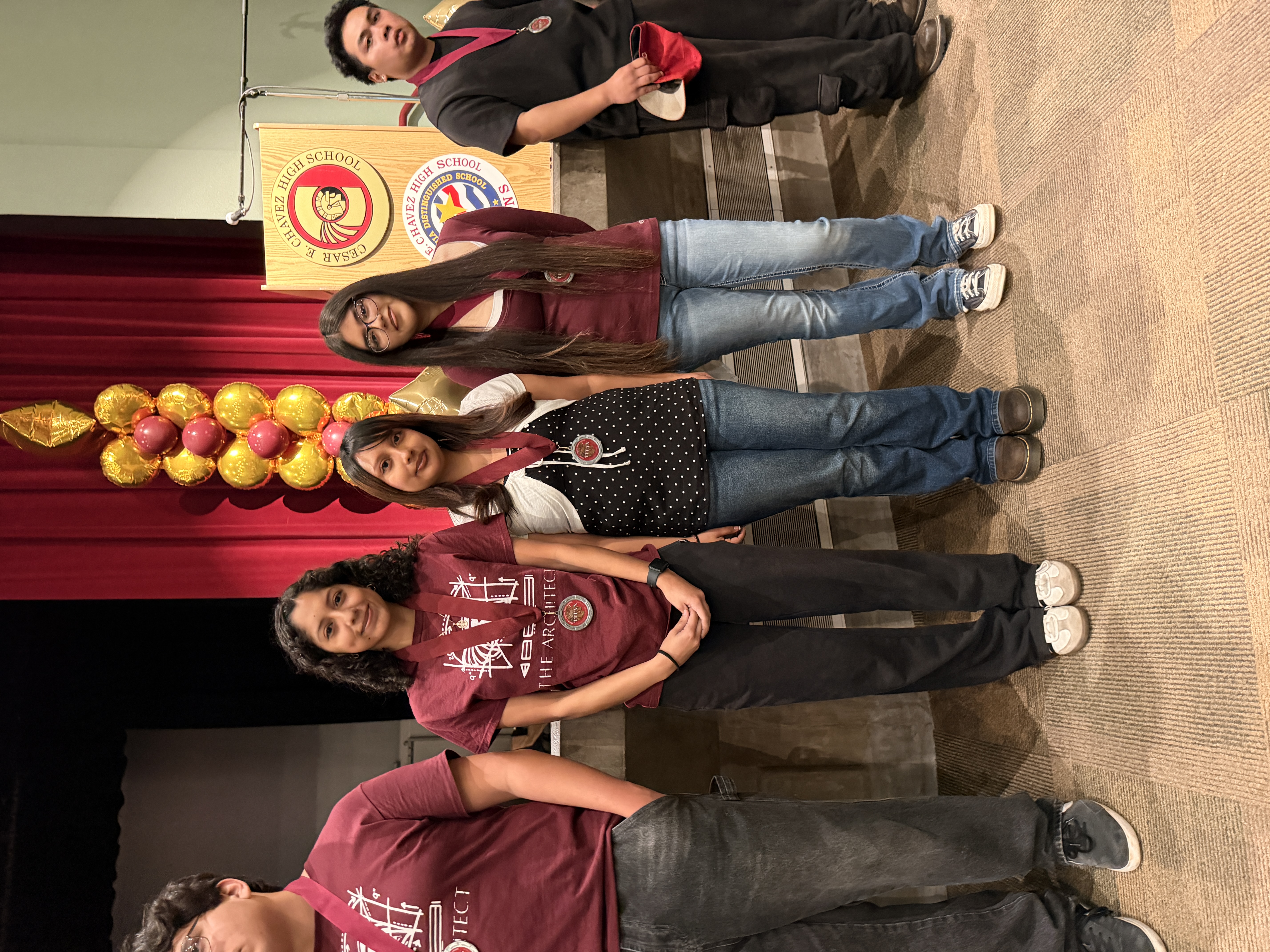 A group of young people, wearing matching maroon shirts, stand in front of a stage with a red curtain and decorative balloons.