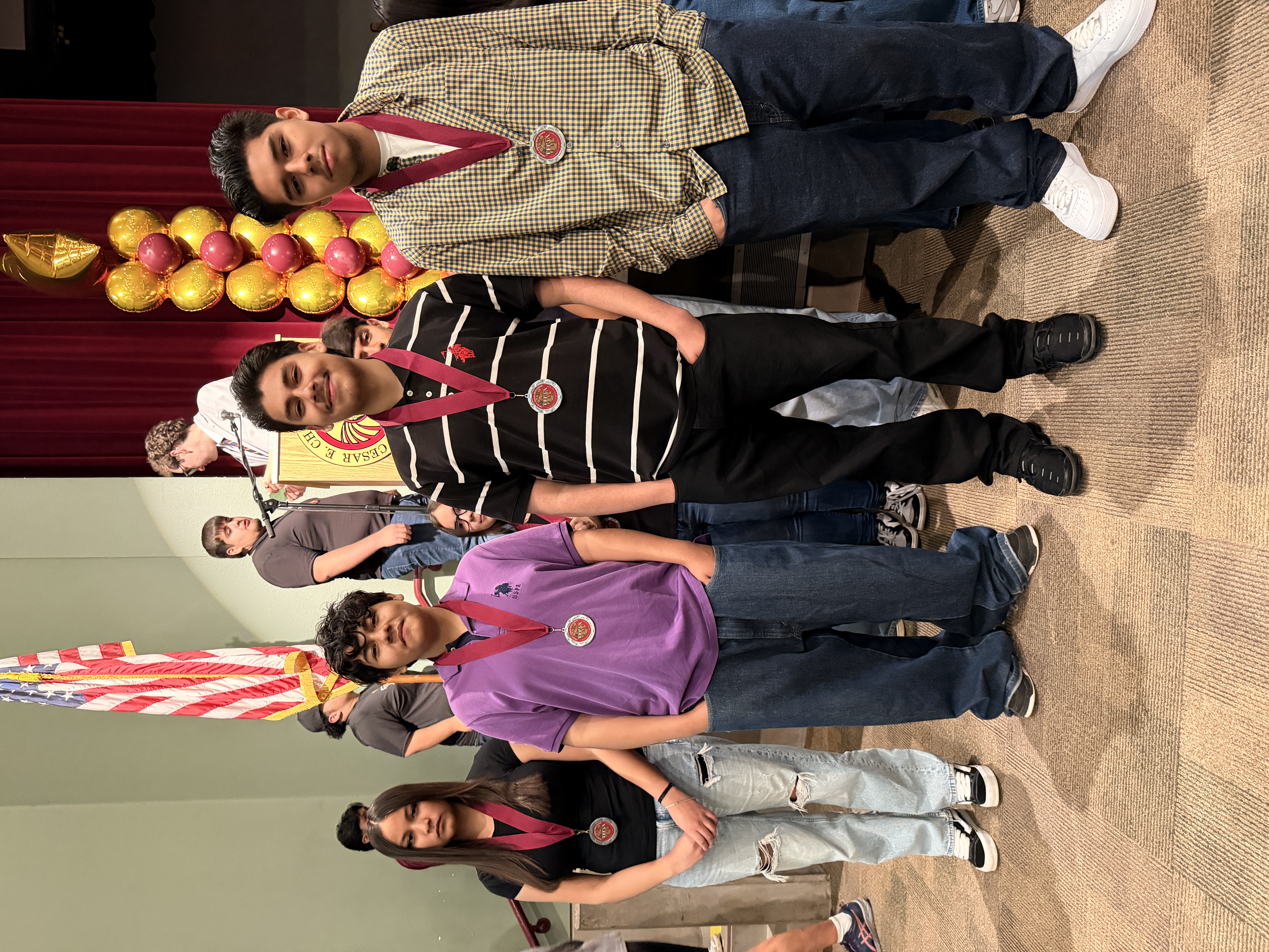 A group of young people, some wearing medals, stand in front of an American flag and a display of gold balloons.