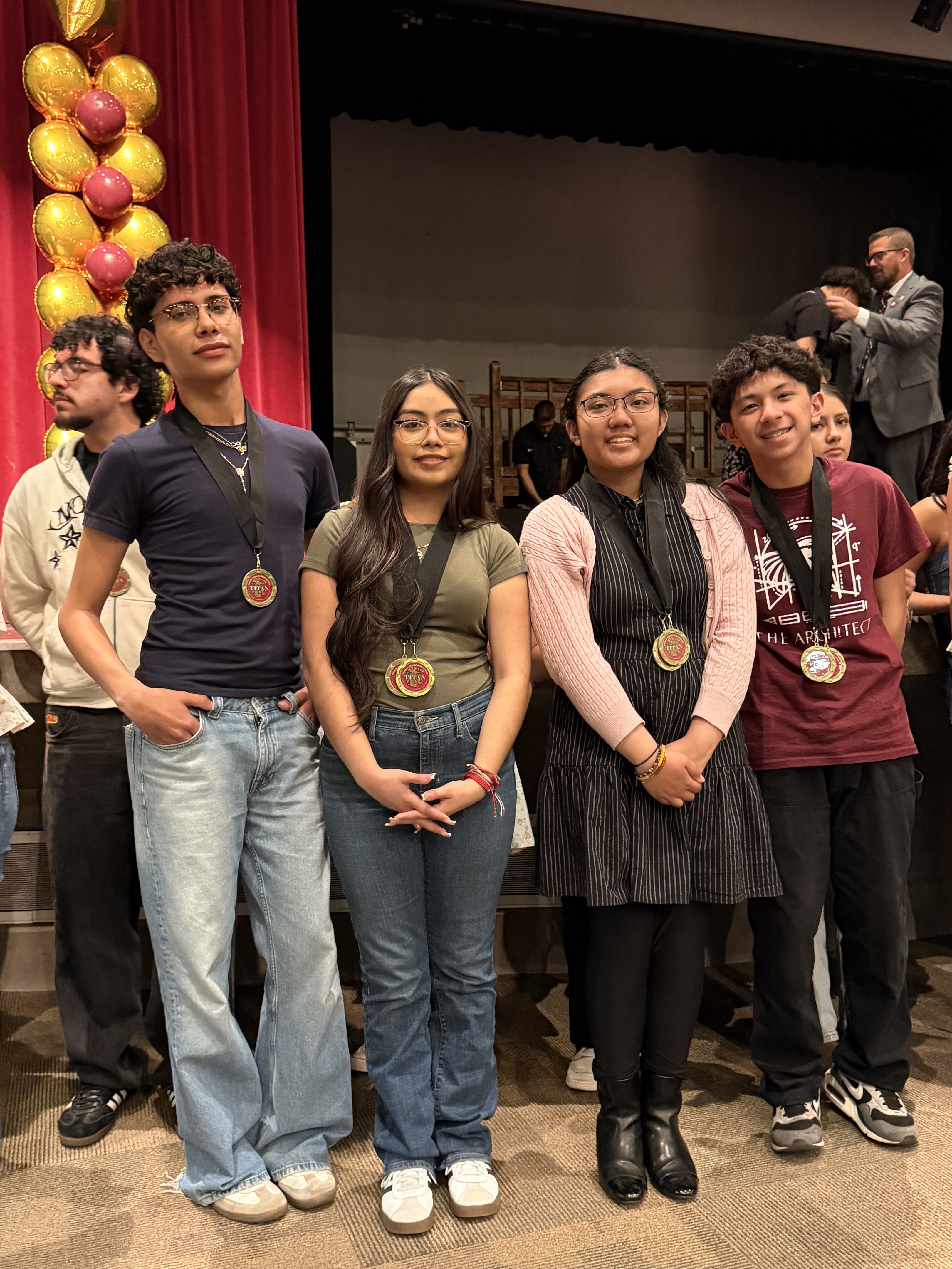 A group of people, including several young adults, stand together in front of a red curtain backdrop, wearing casual clothing and holding medals or awards.