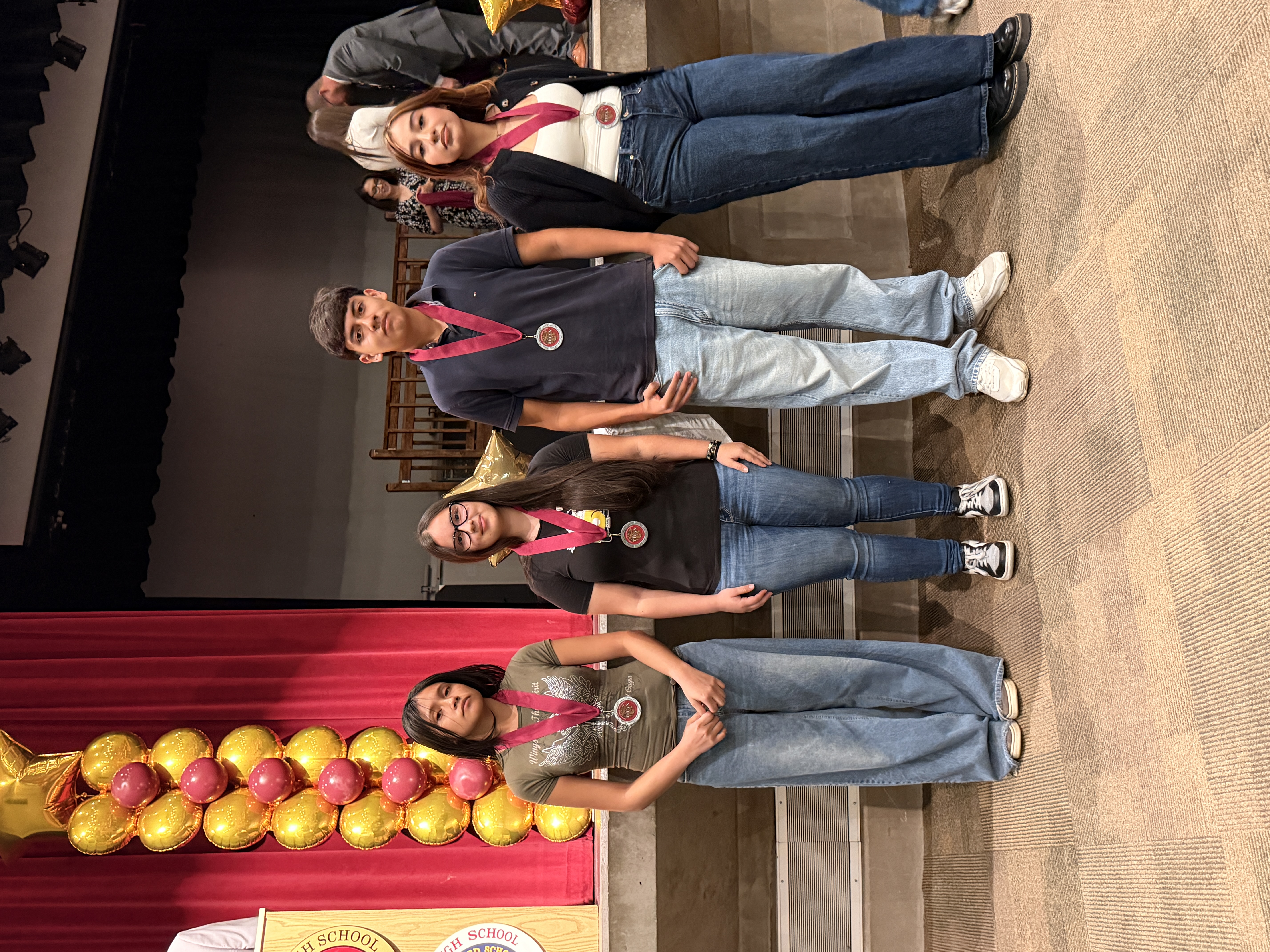 Four individuals, two women and two men, are standing on a stage or platform with a red curtain backdrop and decorative balloons in the background.