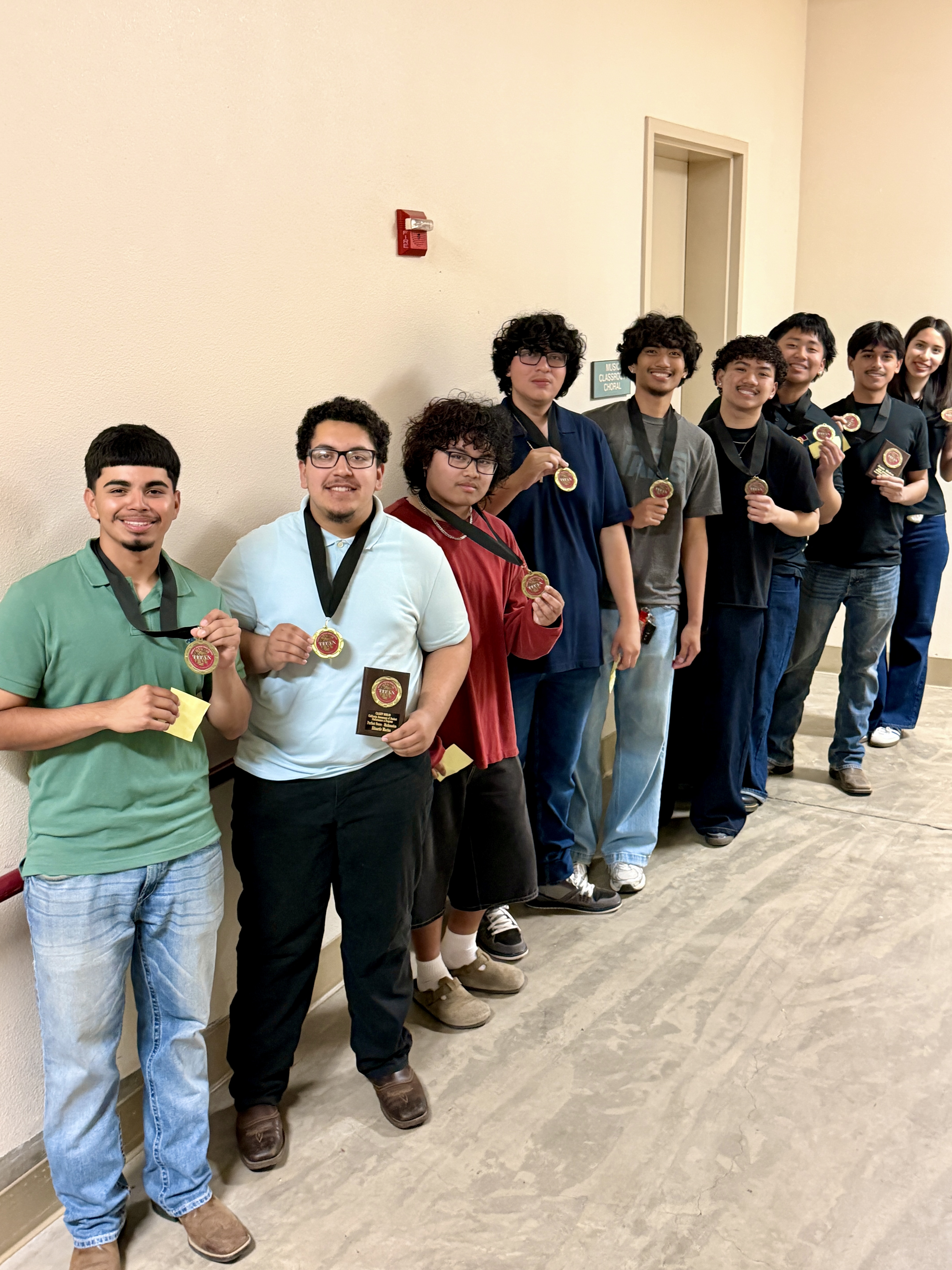 A group of people, mostly men, are standing together in a hallway, some holding medals or trophies, suggesting they have achieved some form of recognition or accomplishment.