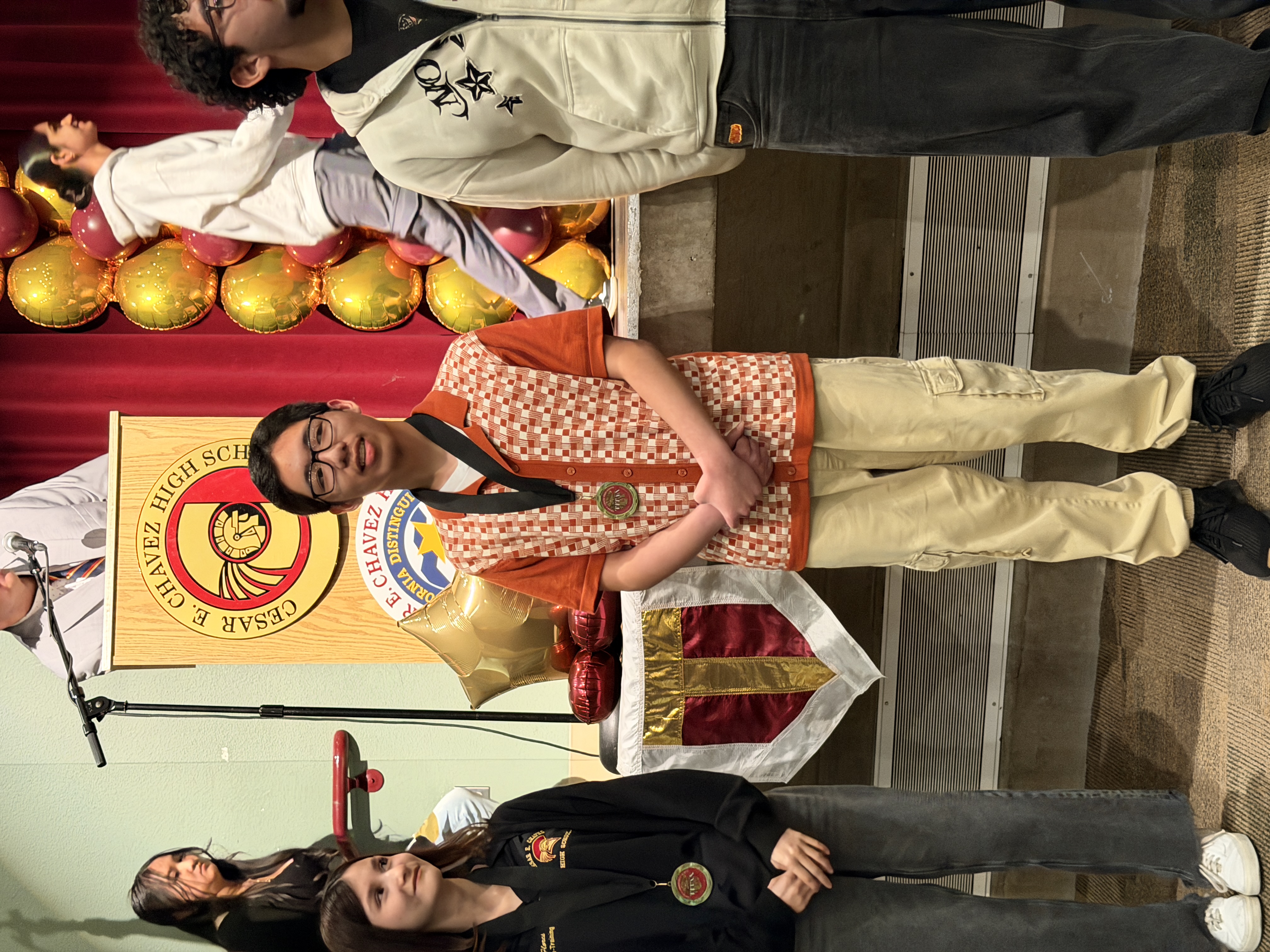 The image shows a group of young people, including a boy wearing a red and white patterned shirt, standing in front of a stage with a banner displaying a sun-like symbol.