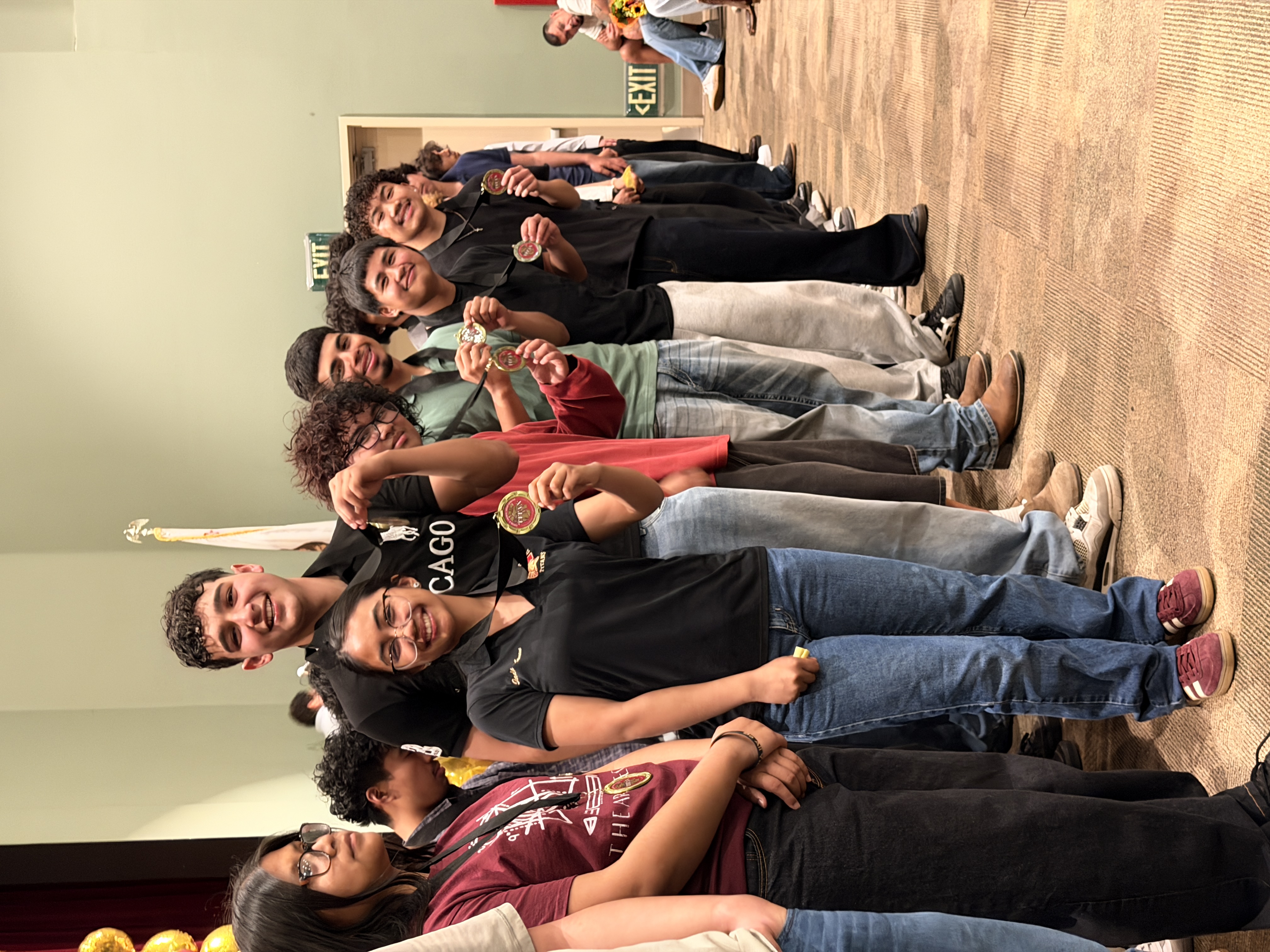 A group of young people, some smiling and others with serious expressions, standing together in what appears to be a hallway or lobby area.
