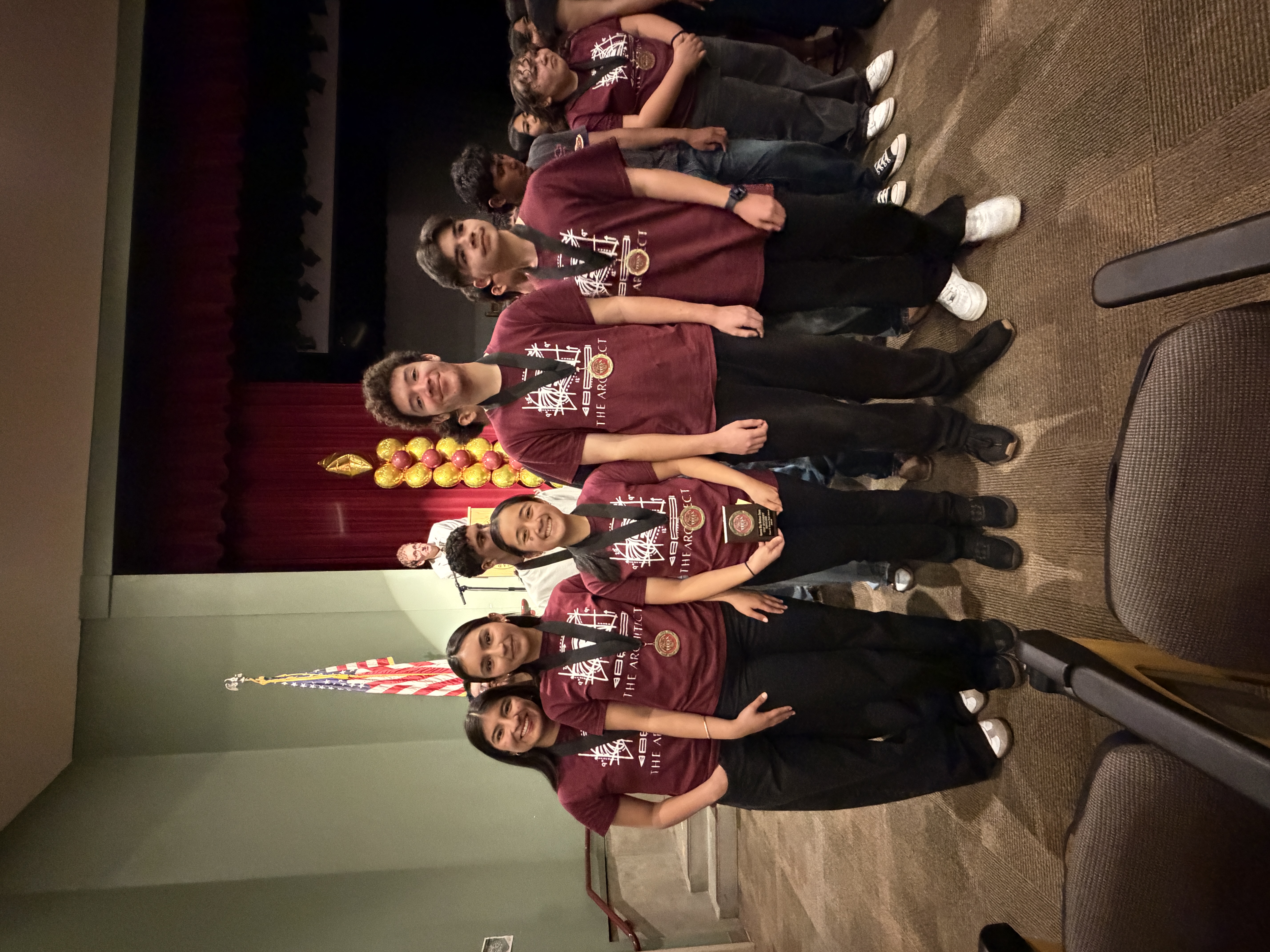 A group of people wearing matching burgundy shirts stand in front of an American flag, with heart-shaped balloons in the background.