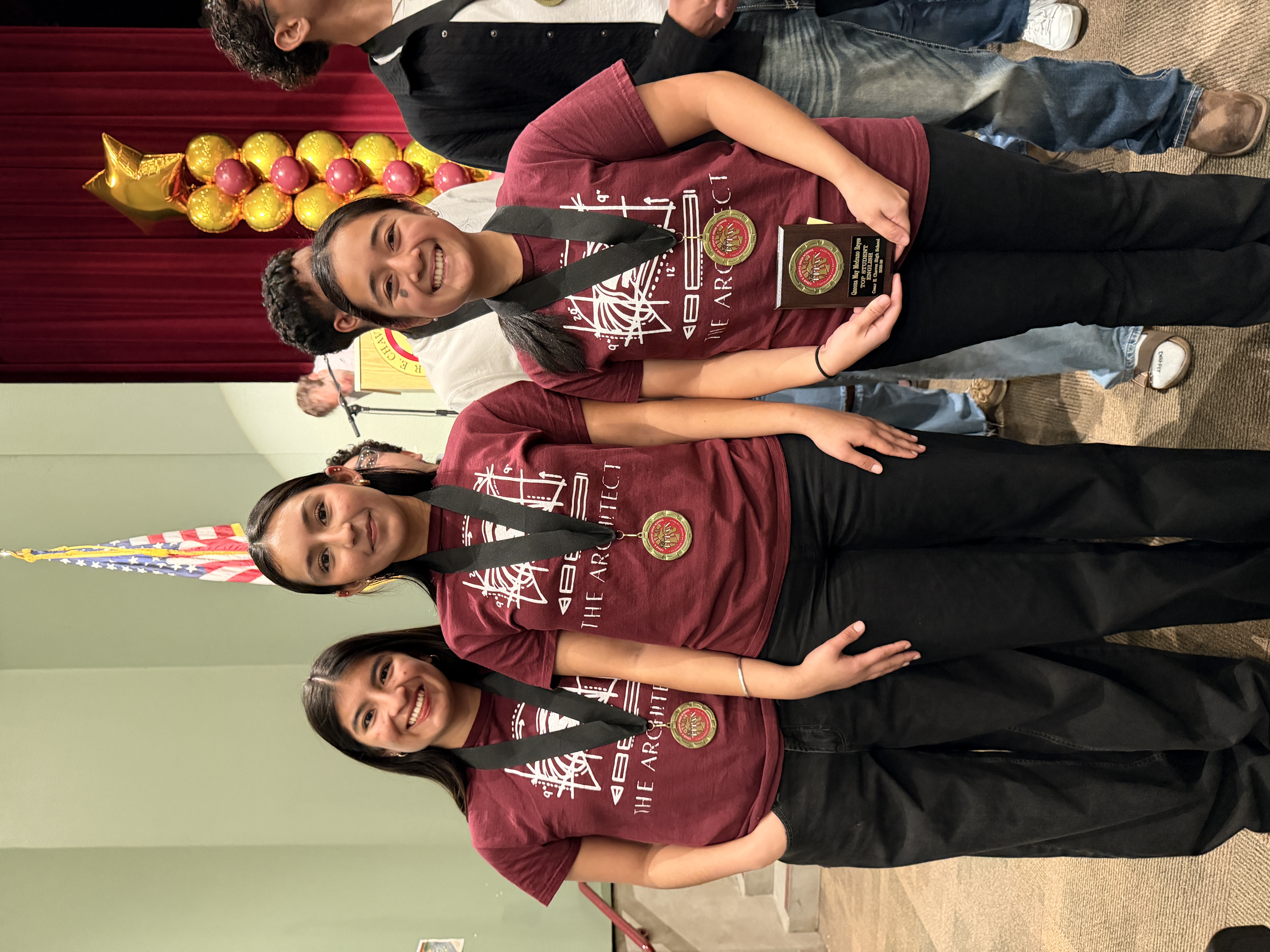 The image shows a group of four people wearing matching burgundy t-shirts with text and logos, standing in front of an American flag and a decorative balloon arch.