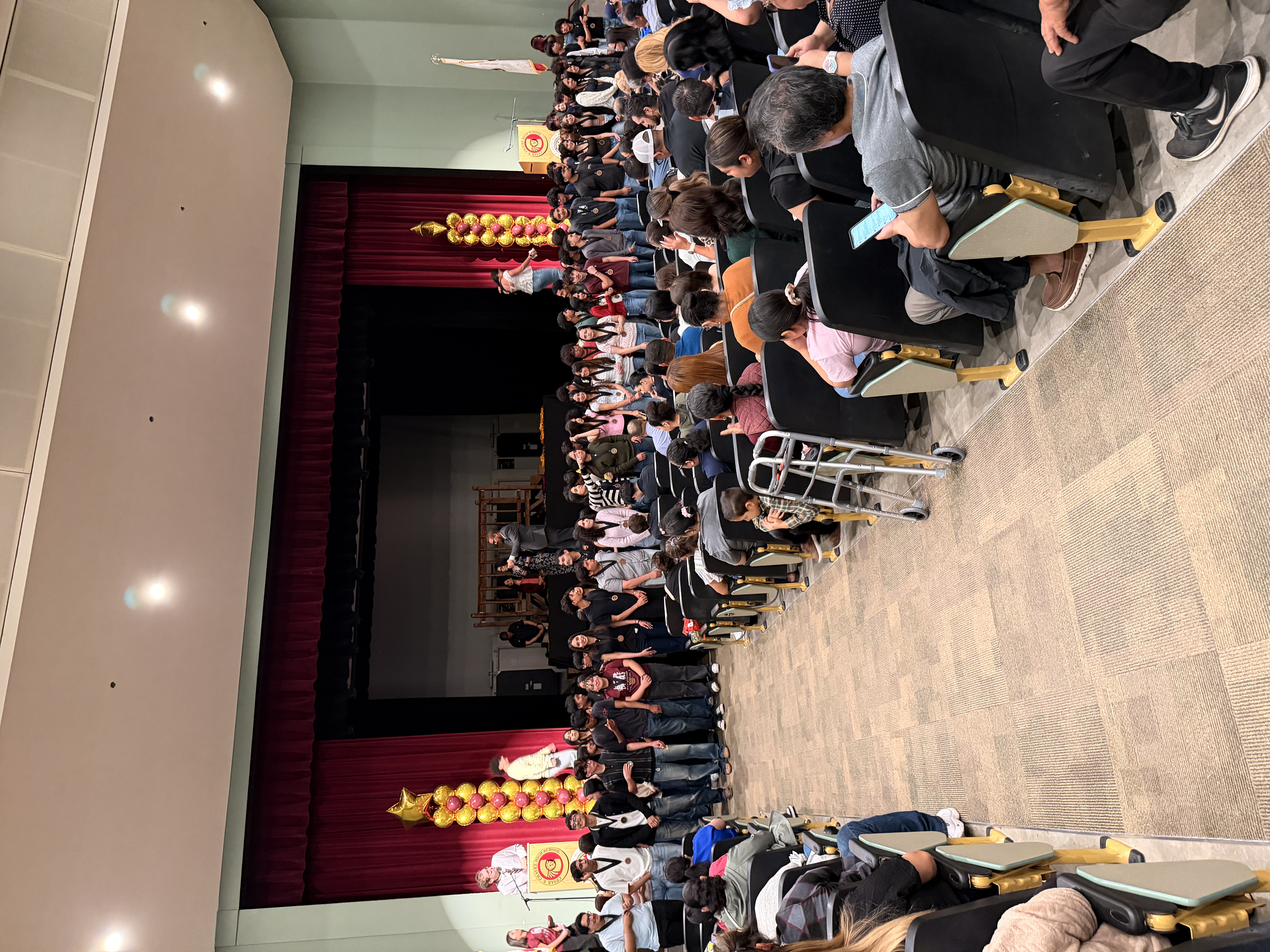 A large crowd of people seated in a theater-like setting, with a stage and decorative elements in the background.