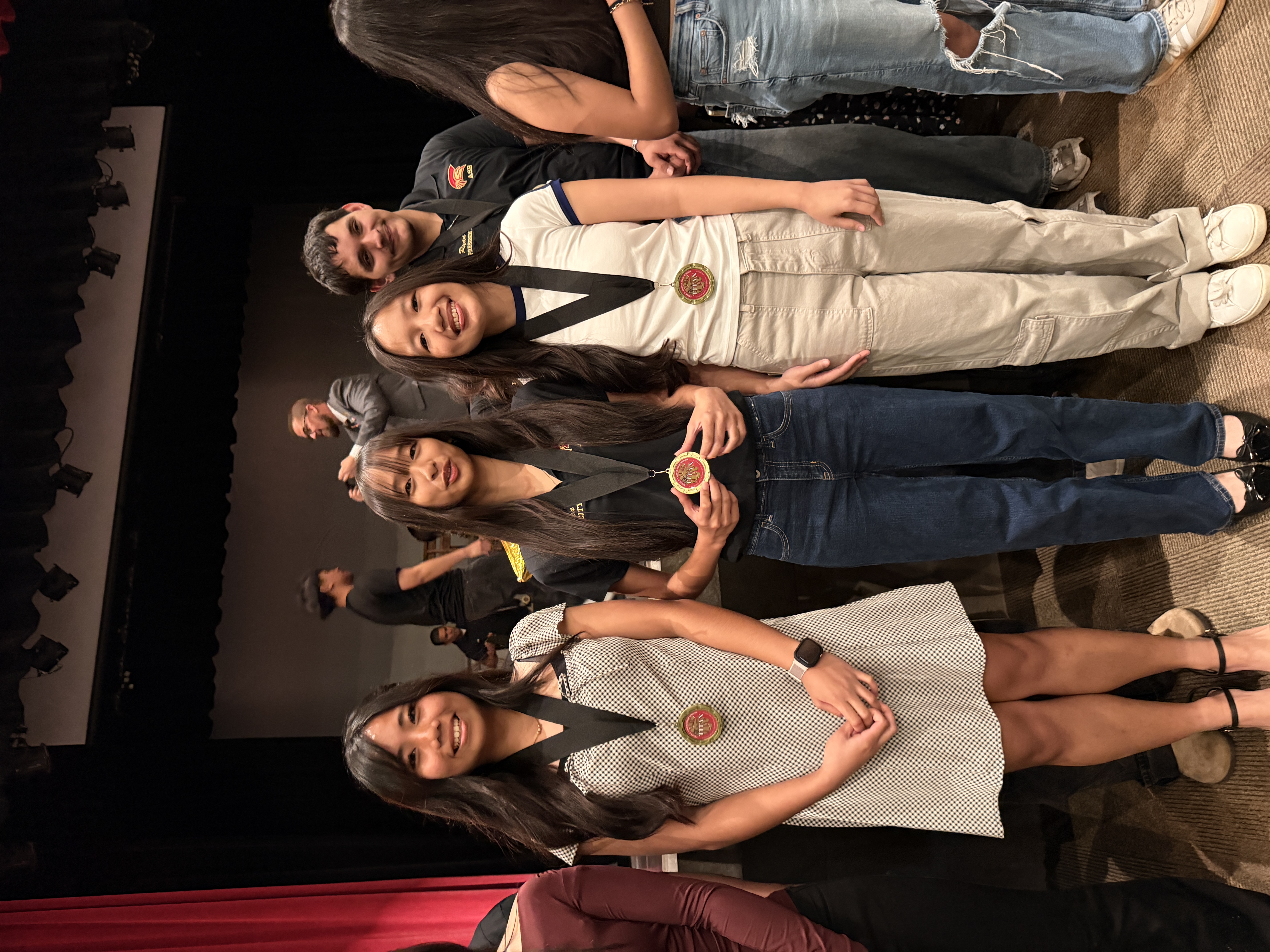 The image shows a group of young women standing on a stage, with a red curtain and stage equipment visible in the background.