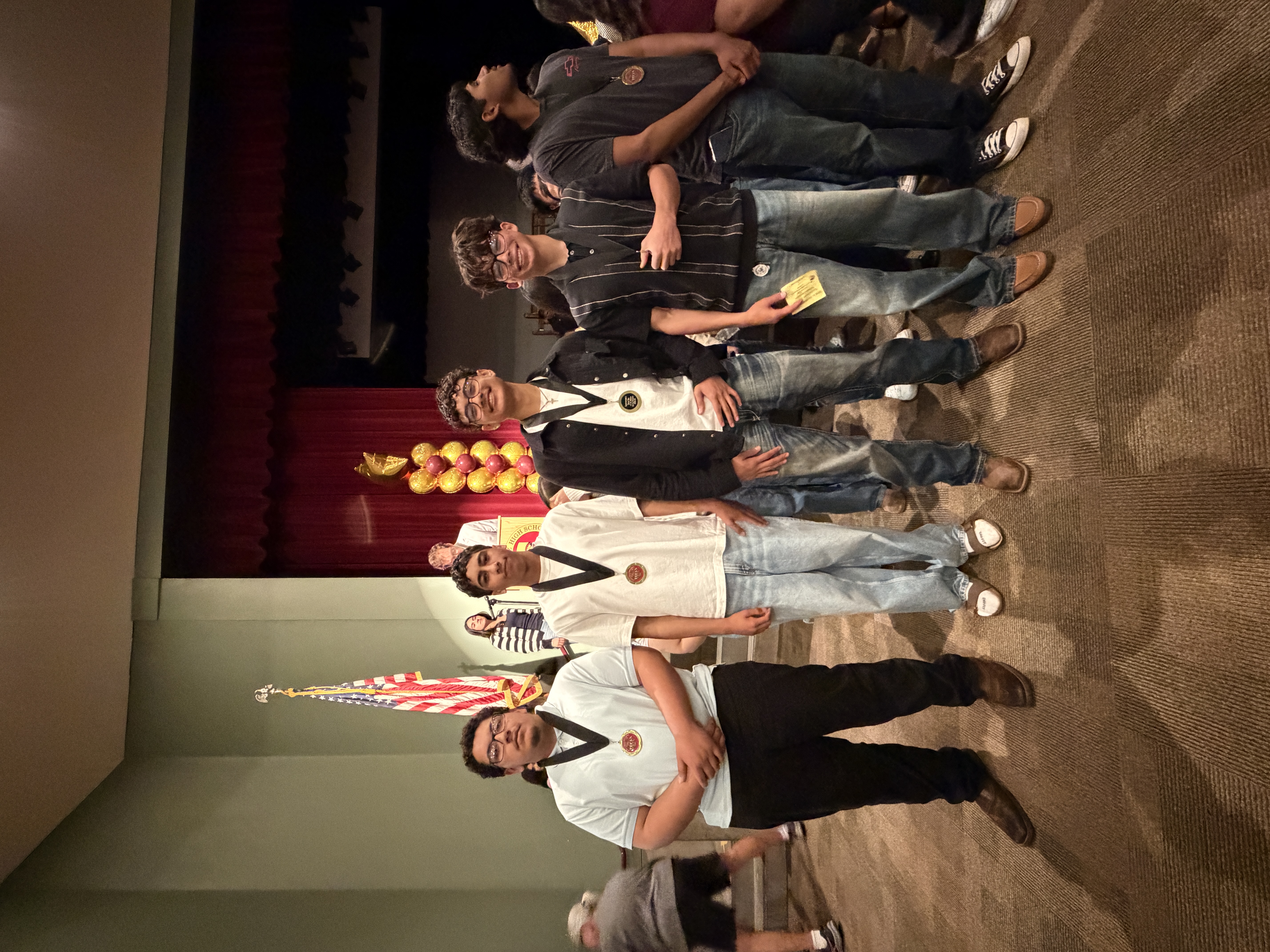 A group of people wearing martial arts uniforms and medals stand in front of a stage with an American flag and decorative balloons in the background.