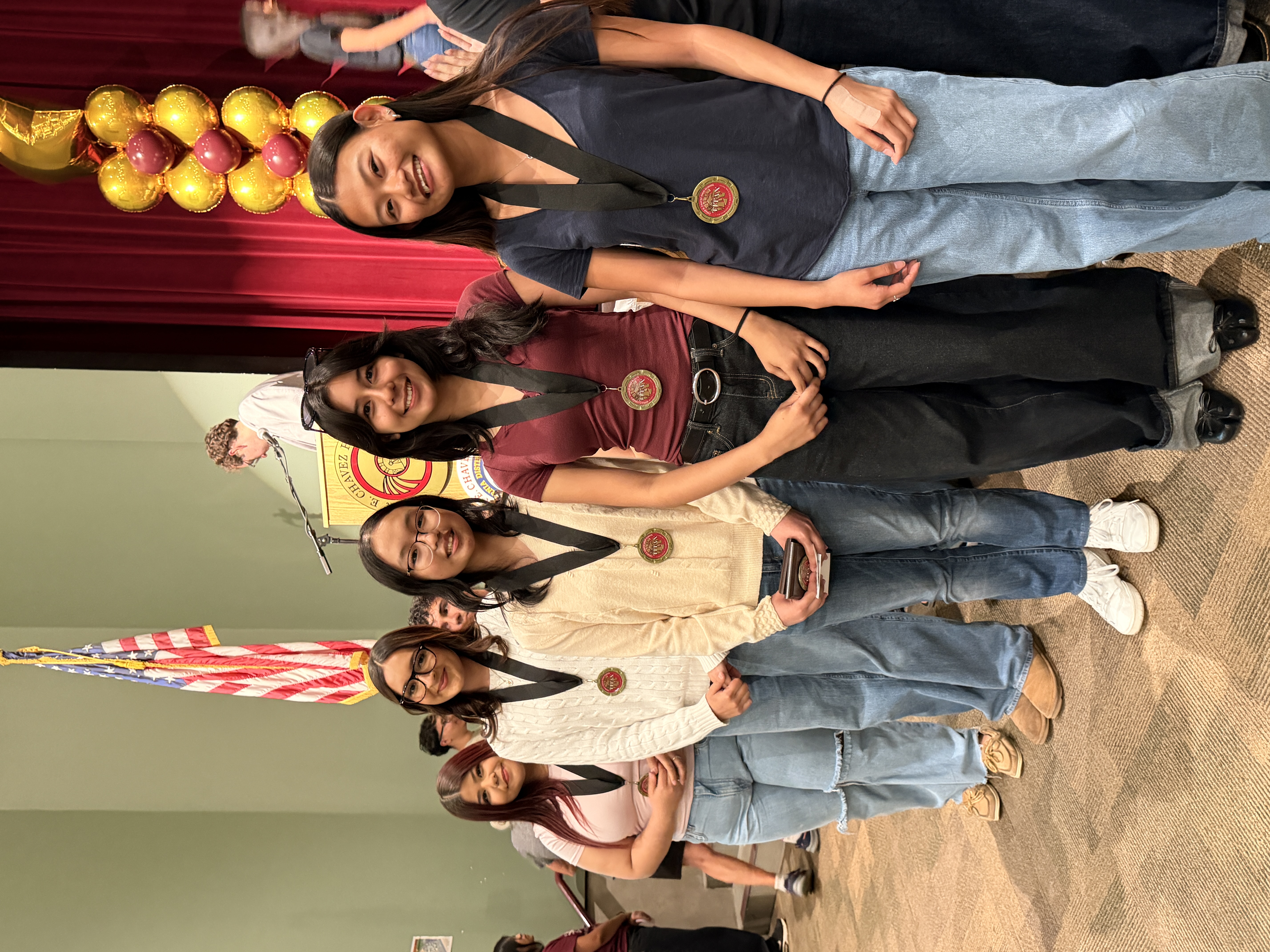 A group of young women in casual attire, some wearing medals, stand in front of a stage with an American flag and decorative balloons in the background.