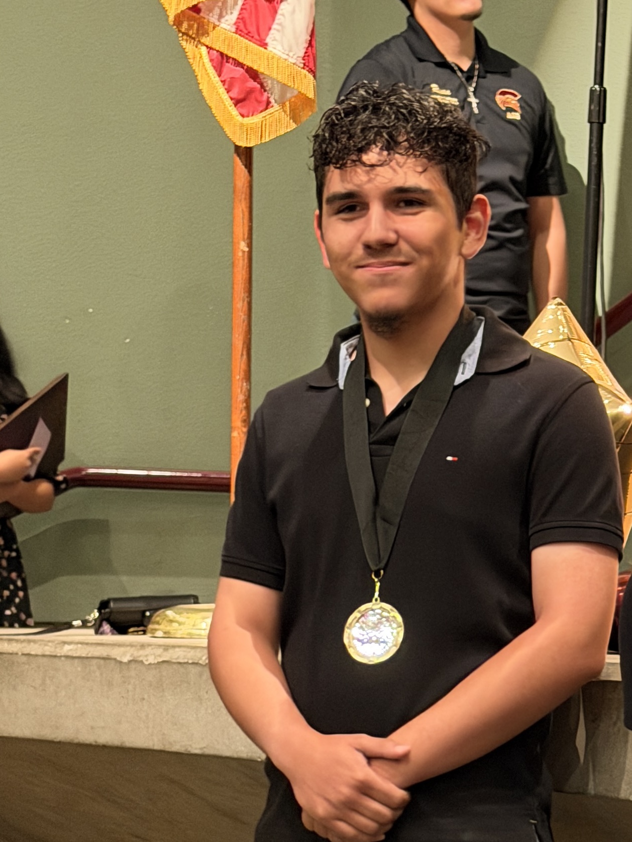 A young man in a black uniform with a medal around his neck stands in front of a colorful flag backdrop.
