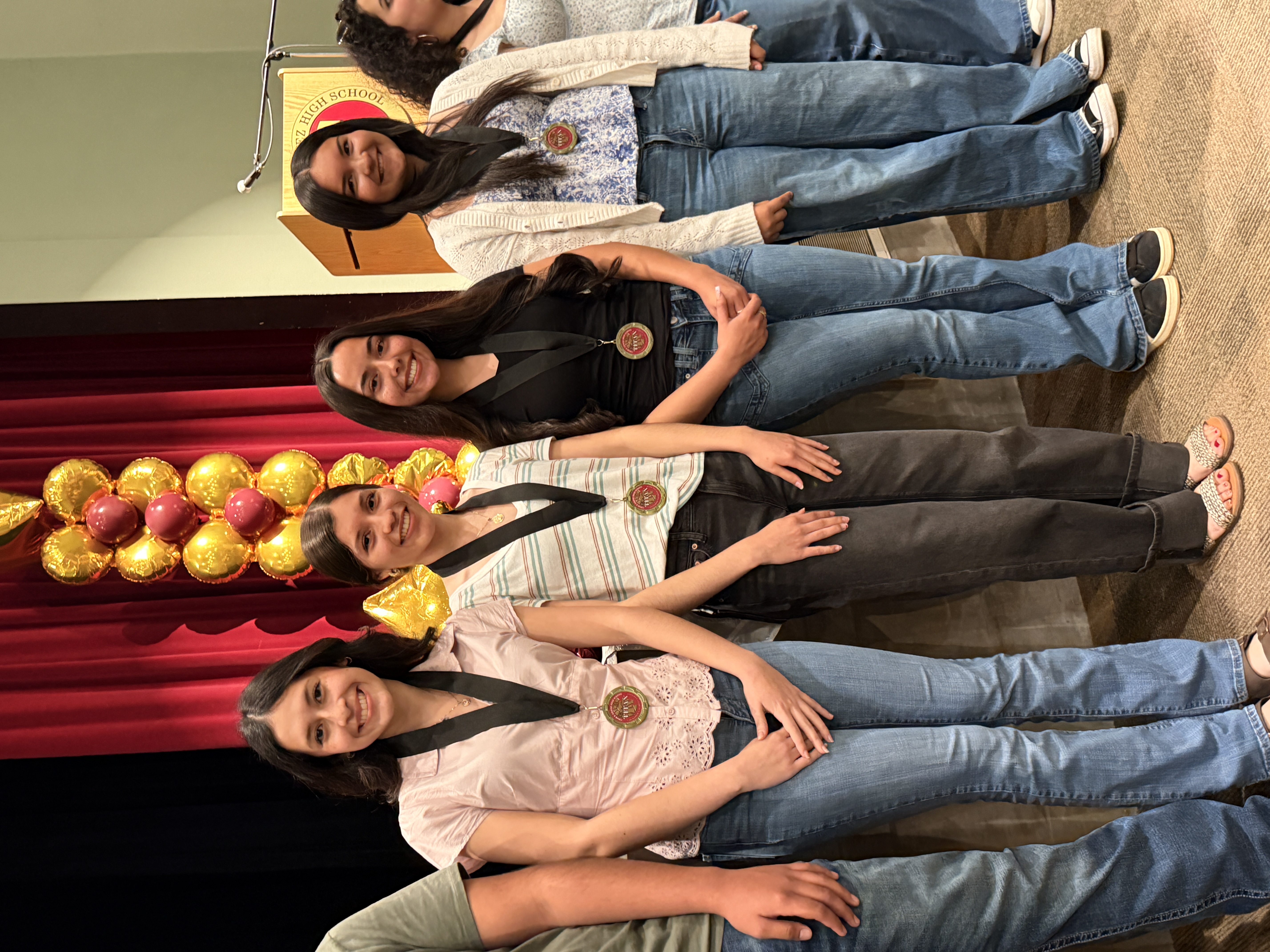 The image shows a group of four young women standing in front of a red curtain backdrop, with a decorative floral arrangement visible in the background.