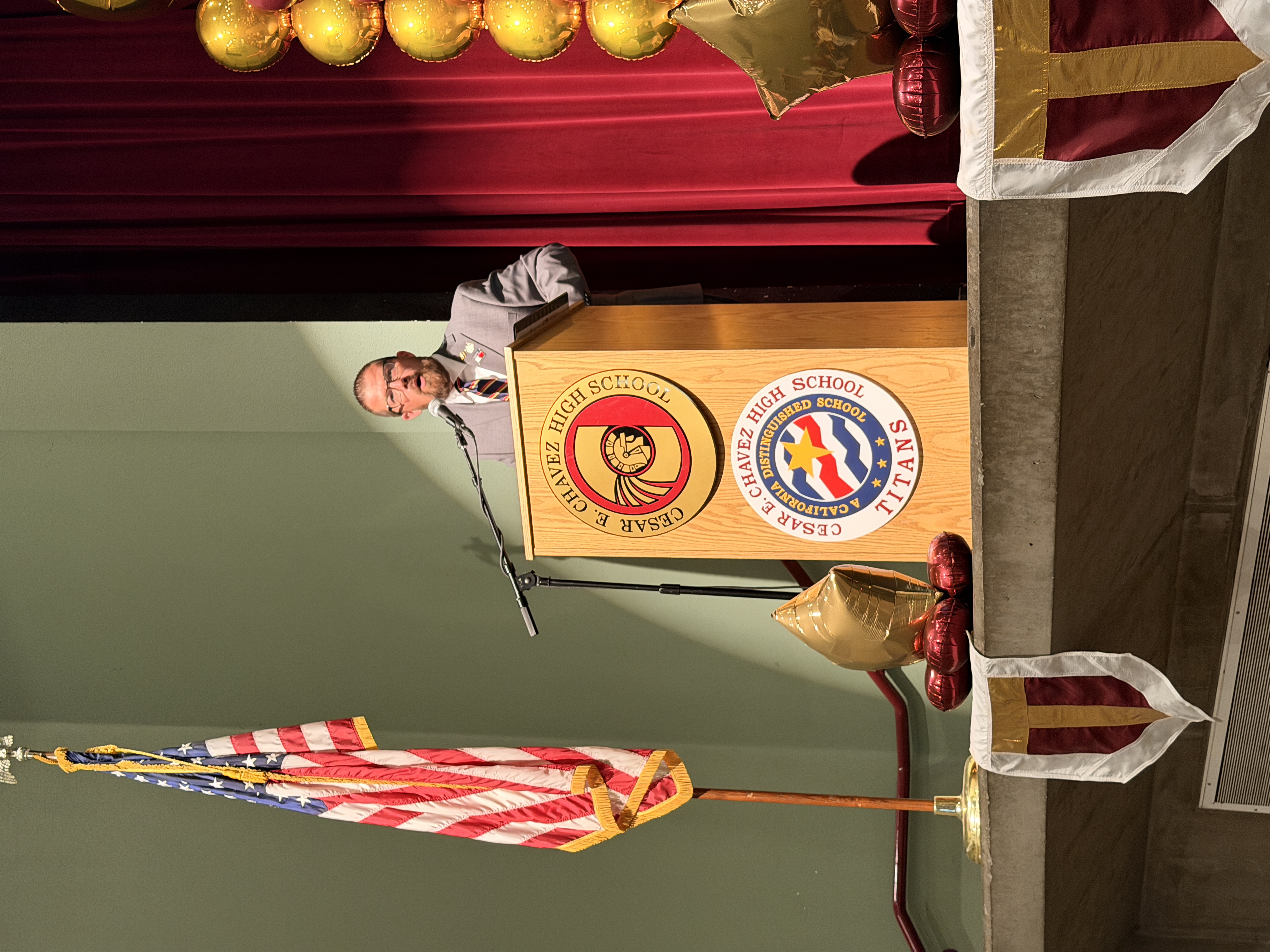 A man in a suit is standing at a podium on a stage, with an American flag and other decorations in the background.